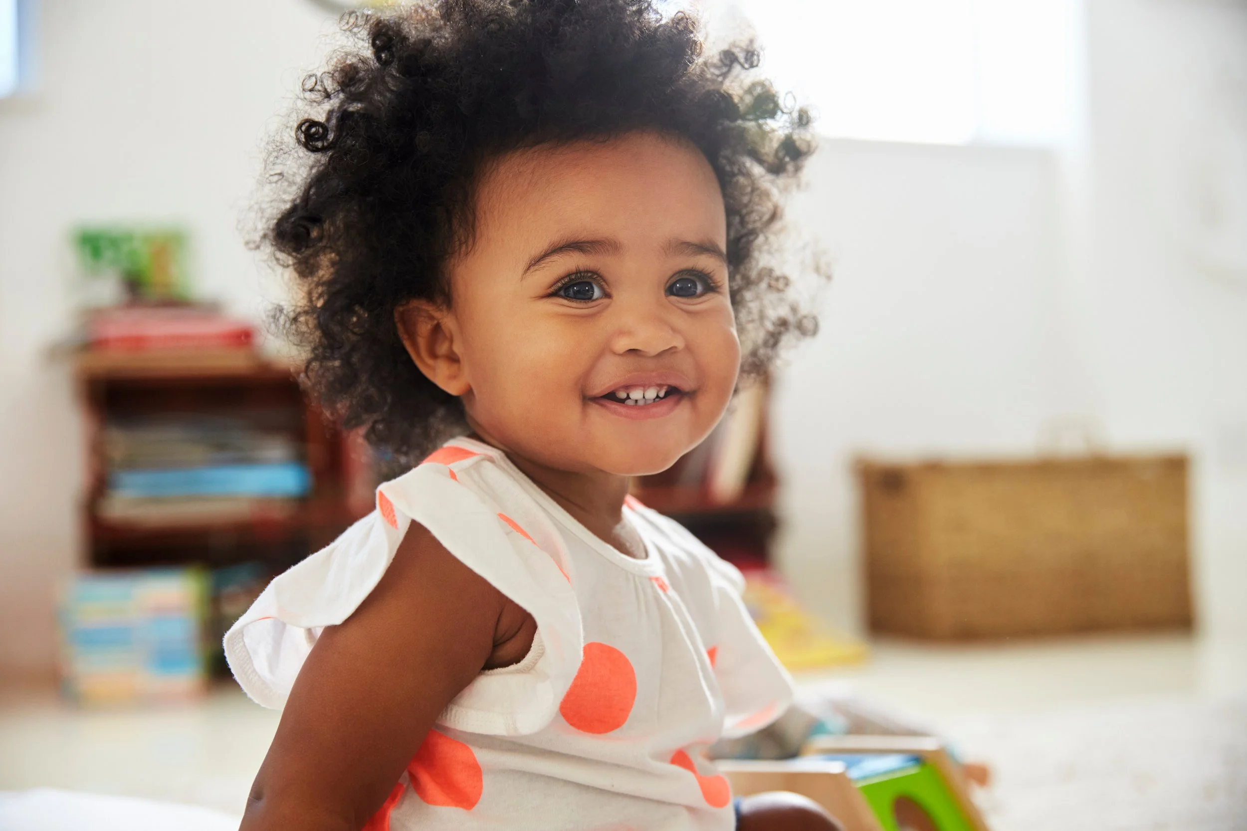 A young girl with curly hair smiling, sitting indoors with a bookshelf and storage basket in the background.