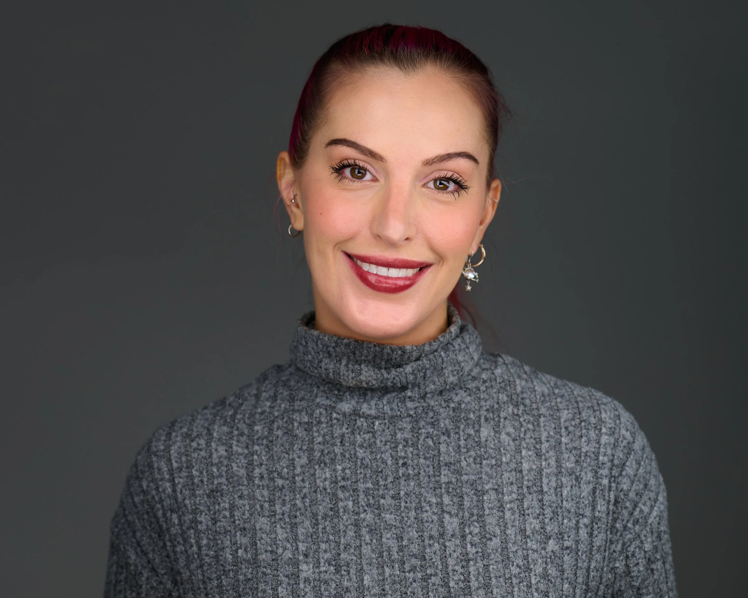Close-up portrait of a young woman with long wavy brown hair, brown eyes, and a gentle smile, wearing a beige blazer and a white collared shirt, against a dark background.