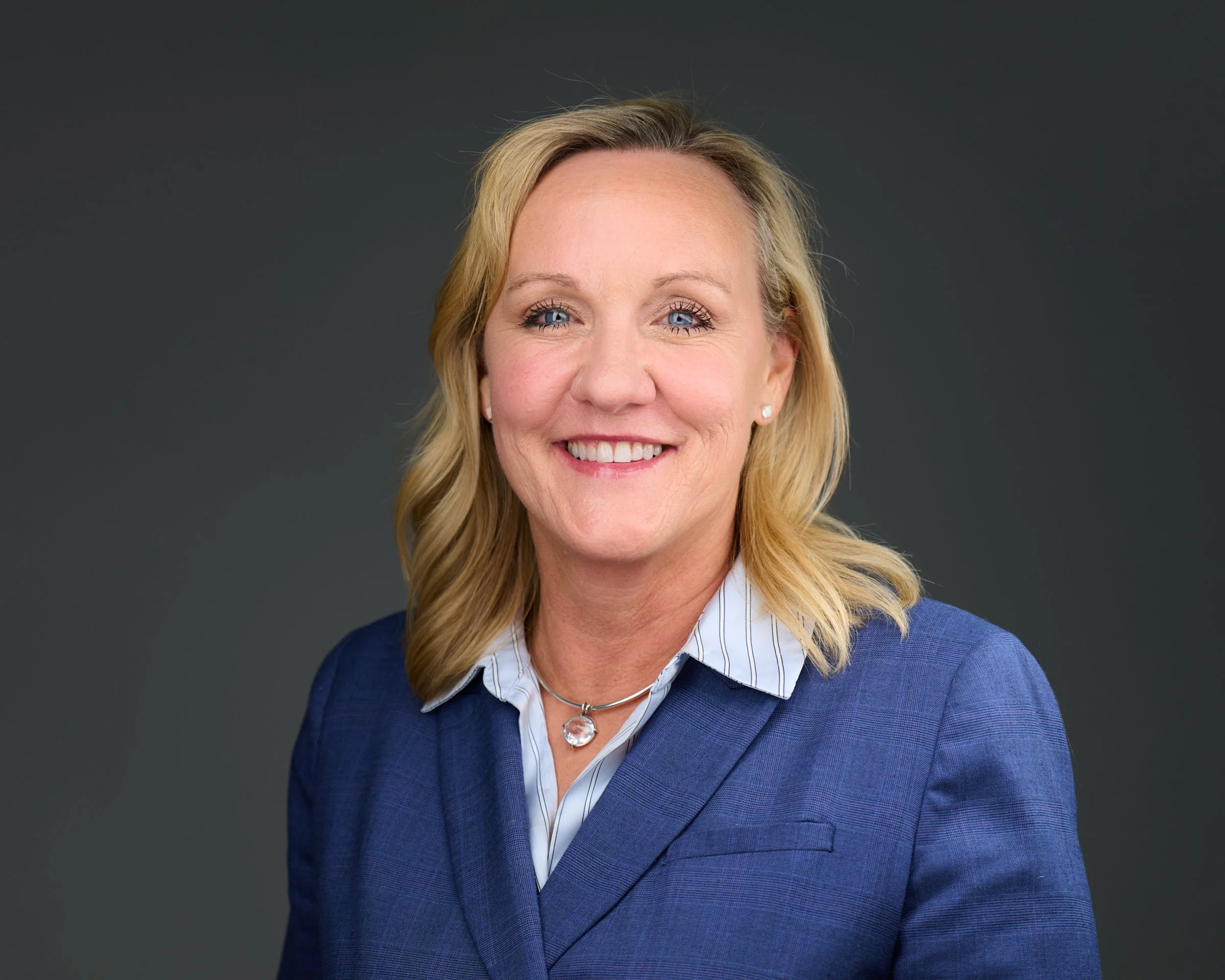 A woman with blonde hair wearing a blue blazer and earrings, smiling against a black background.