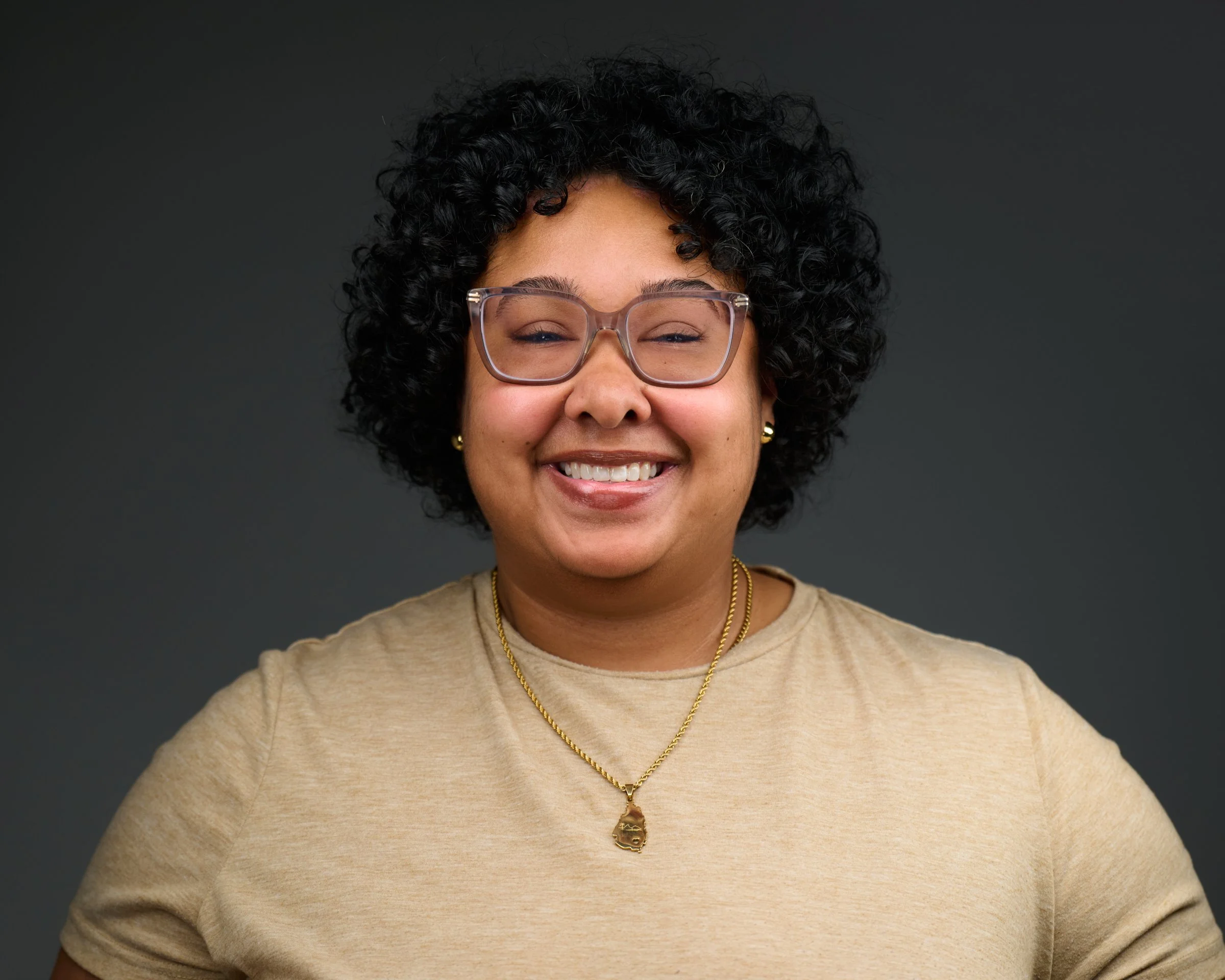 Close-up of a woman with dark curly hair, wearing glasses, gold jewelry, and a black top, smiling at the camera.