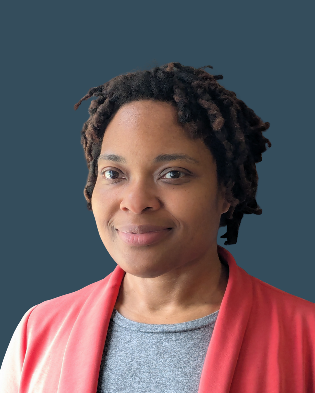 A woman with dark skin and dreadlocks smiling, wearing a floral headscarf, a pink shirt, and a blue sweater, against a dark background.