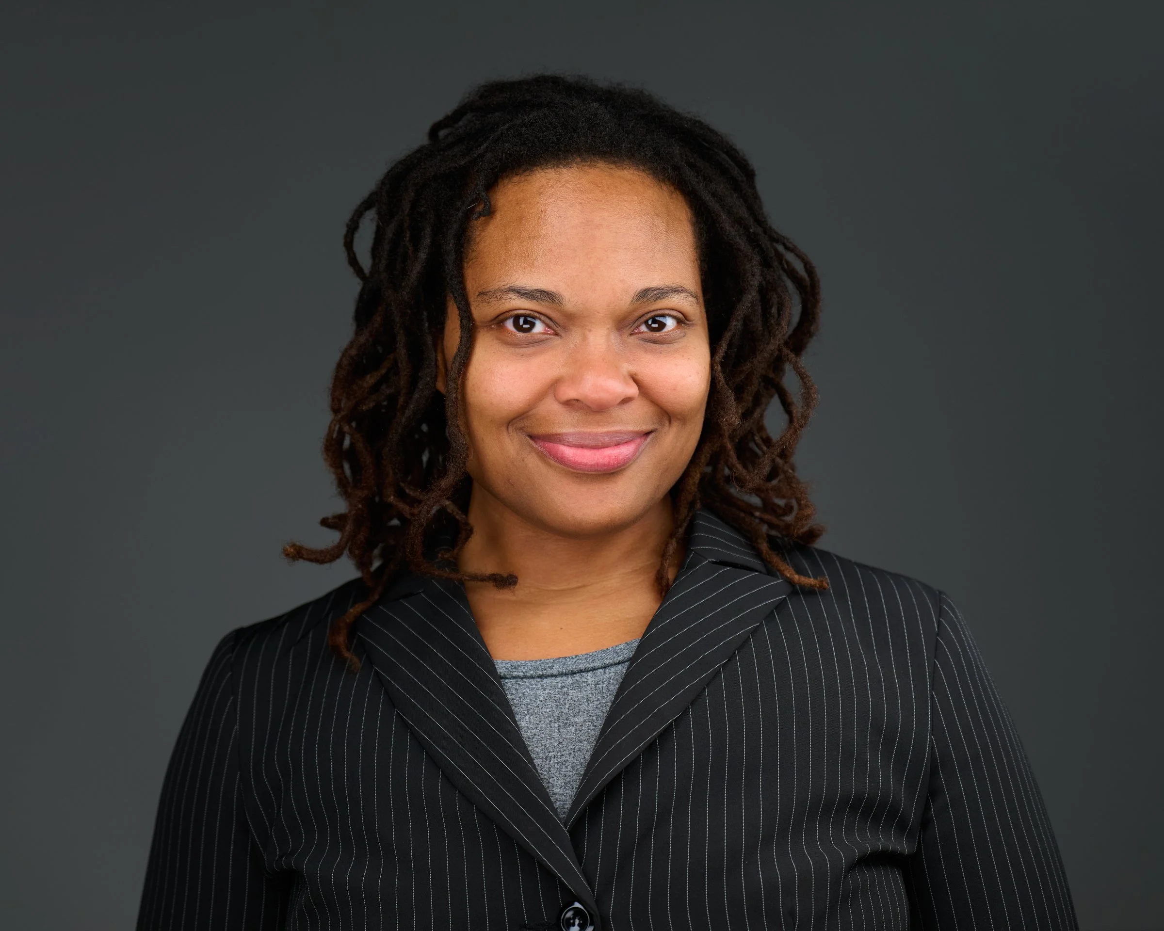 A woman with dark skin and dreadlocks smiling, wearing a floral headscarf, a pink shirt, and a blue sweater, against a dark background.