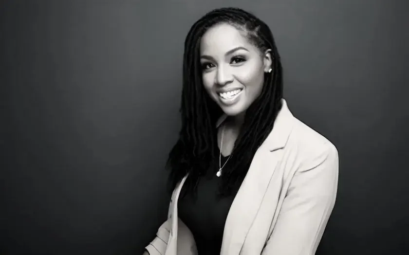 Black and white portrait of a smiling woman with dreadlocks, wearing a light blazer and a necklace, standing against a plain dark background.