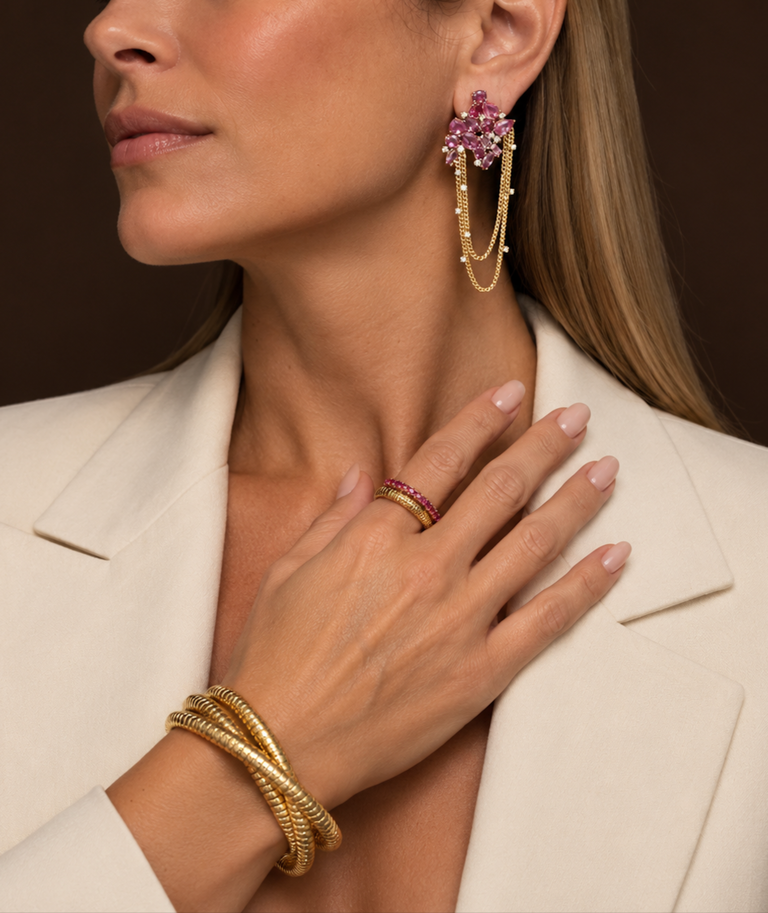 Close-up of a woman wearing a white blazer, large pink gemstone and gold dangling earring, multiple gold bangles, a pink gemstone ring, and a blonde hairstyle. Her lips are slightly parted and her hand is resting on her chest.