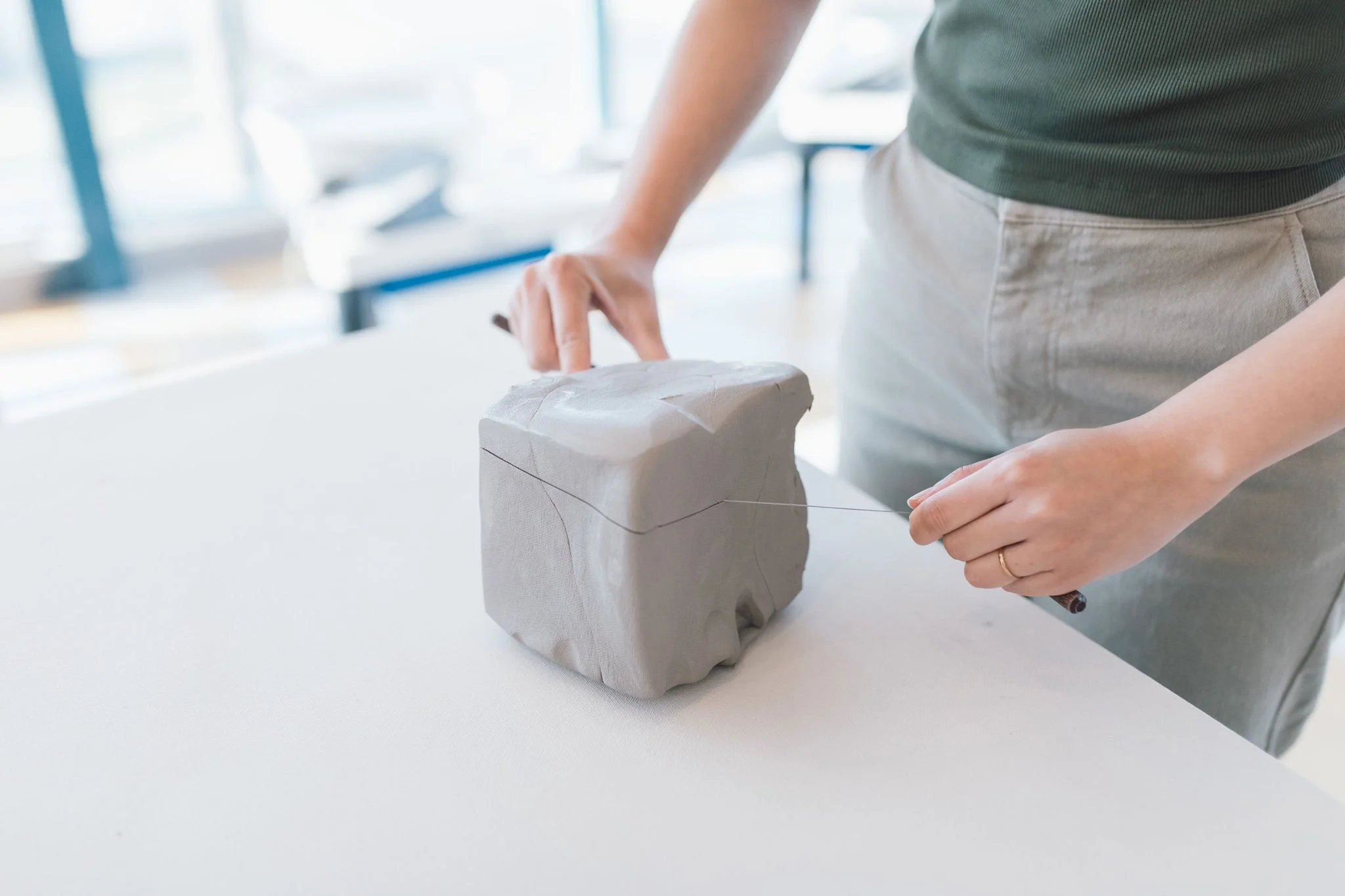 A woman skillfully shaping a grey, square clay block  on a table with a cutting tool.