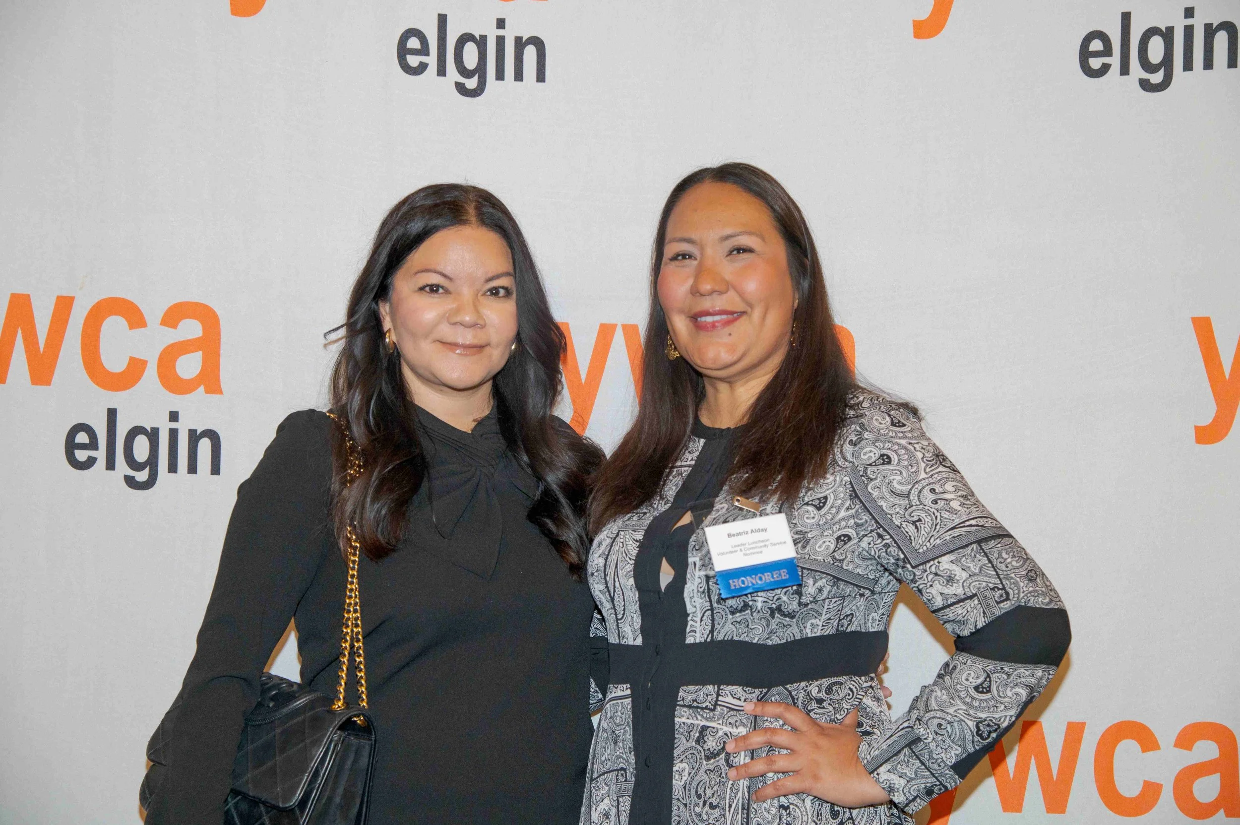 Two women standing side by side, smiling for the camera at an event with a backdrop that says 'elgin'. The woman on the left has long dark hair, is wearing a black outfit with a gold chain shoulder bag, and earrings. The woman on the right has long d