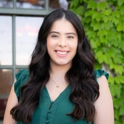 A woman smiling, dressed in a colorful traditional Mexican dress with ruffled ribbons and jewelry, against a solid blue background.