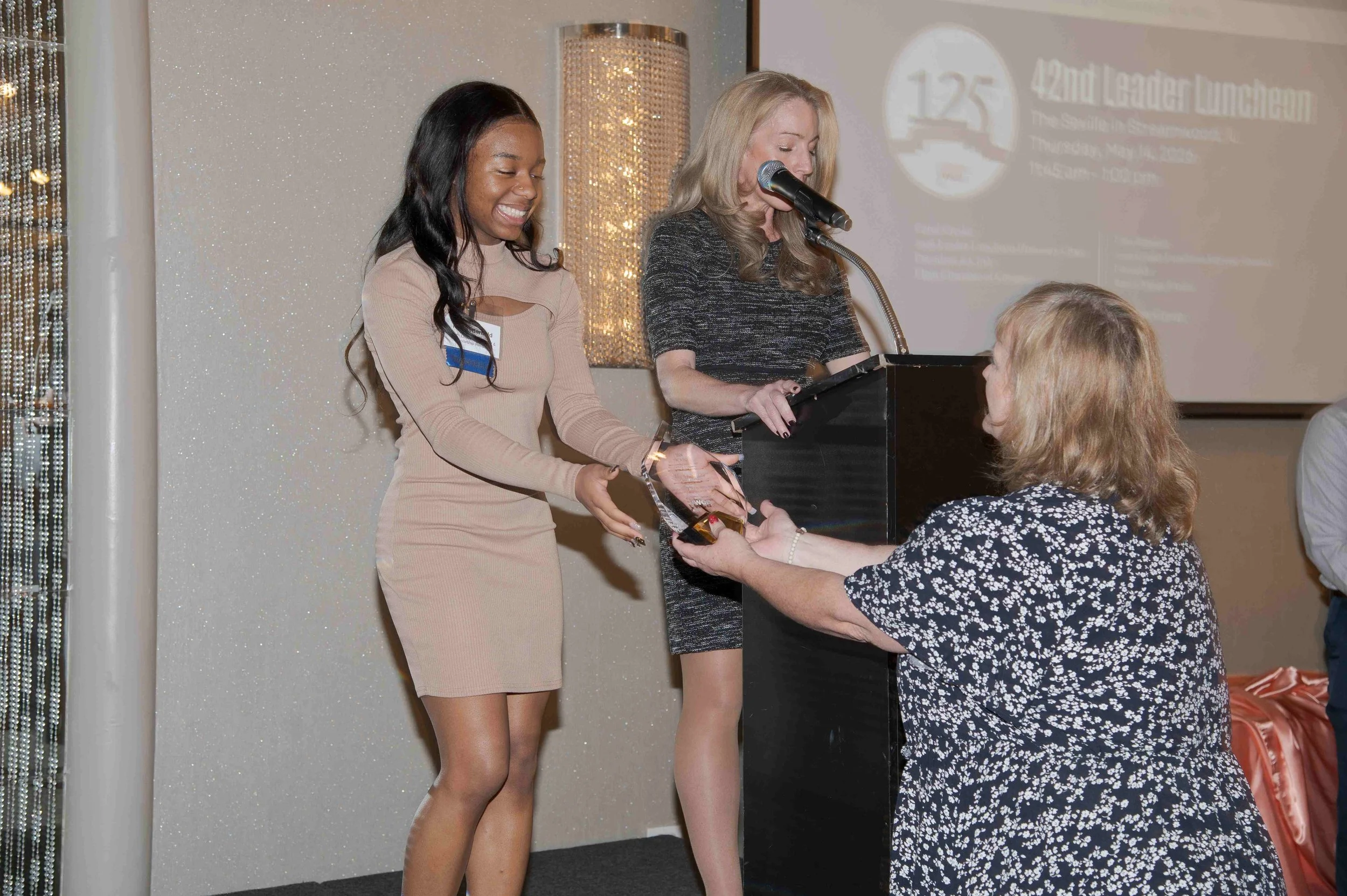 Two women are on stage at an awards event; the woman on the left is smiling as she receives an award from the woman on the right, who is speaking into a microphone. The background features a projected logo and decorative curtains.
