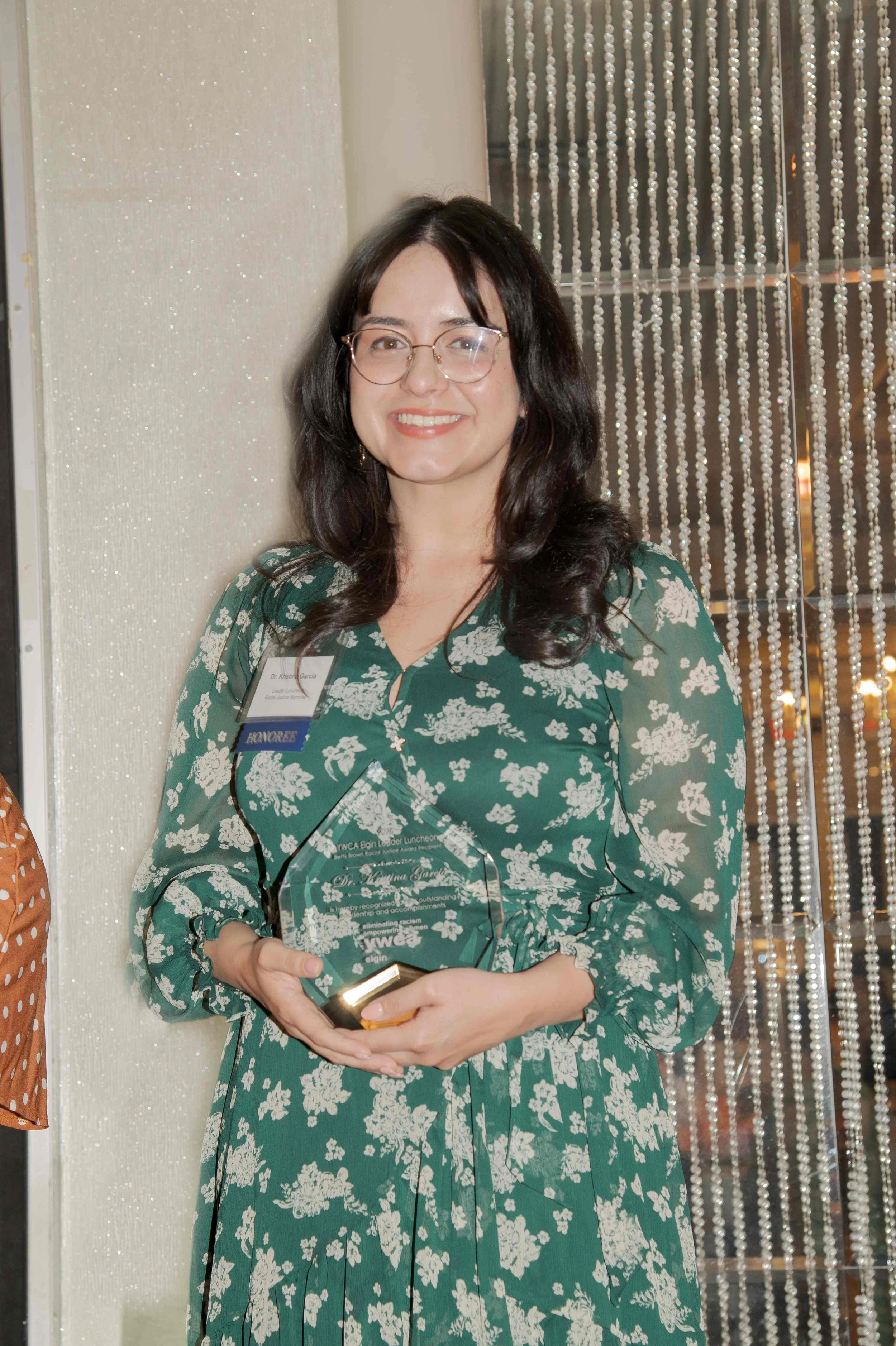 A woman with dark hair and glasses smiling while holding a glass award, wearing a green dress with white floral pattern, standing in front of a beaded curtain at an indoor event.