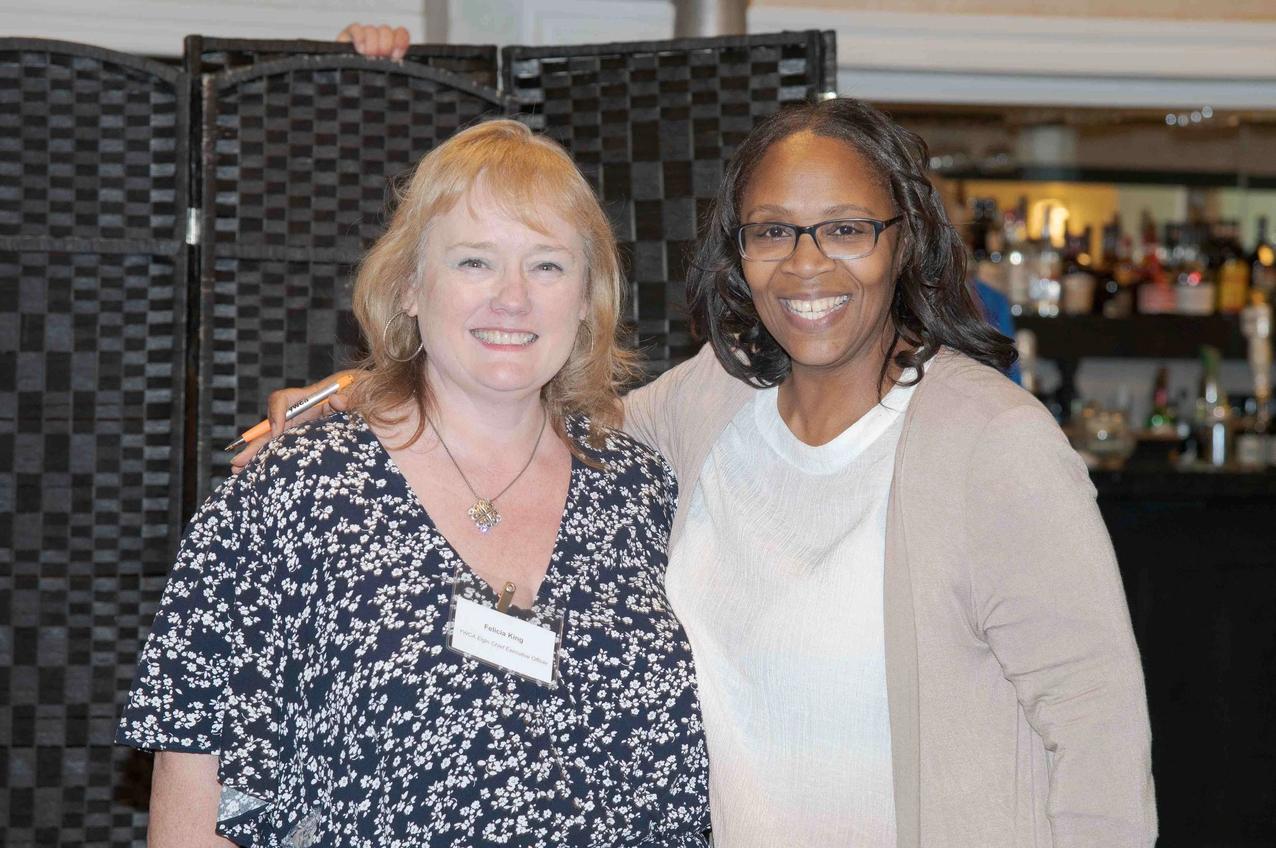 Two women smiling, standing close together at a social event; the woman on the left has red hair, wears a black and white floral top, and has a name tag; the woman on the right has dark hair, wears glasses, and a beige blazer over a white top. They a