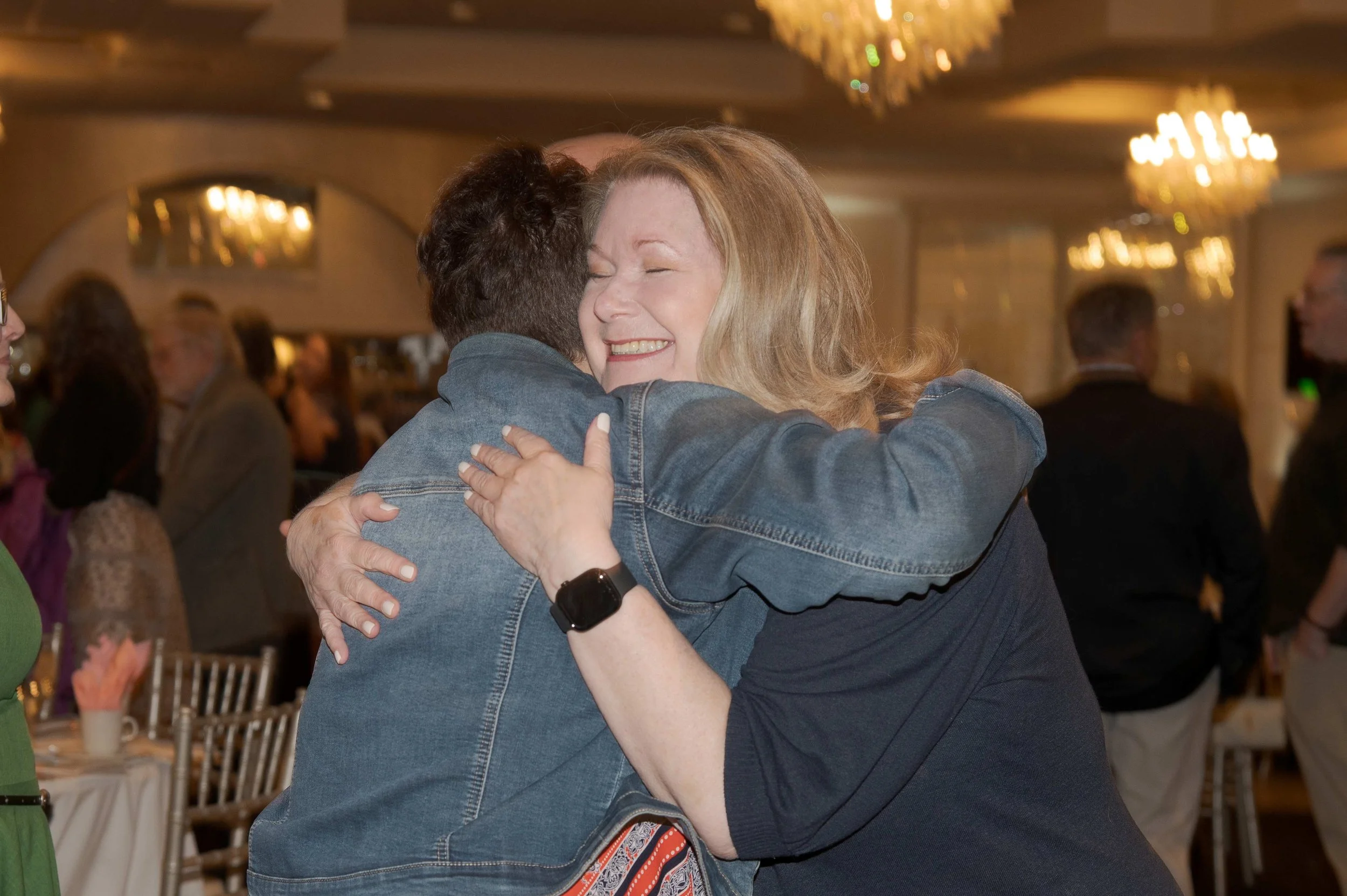 Two women hugging, smiling, and enjoying a joyful moment in a celebration or event setting.