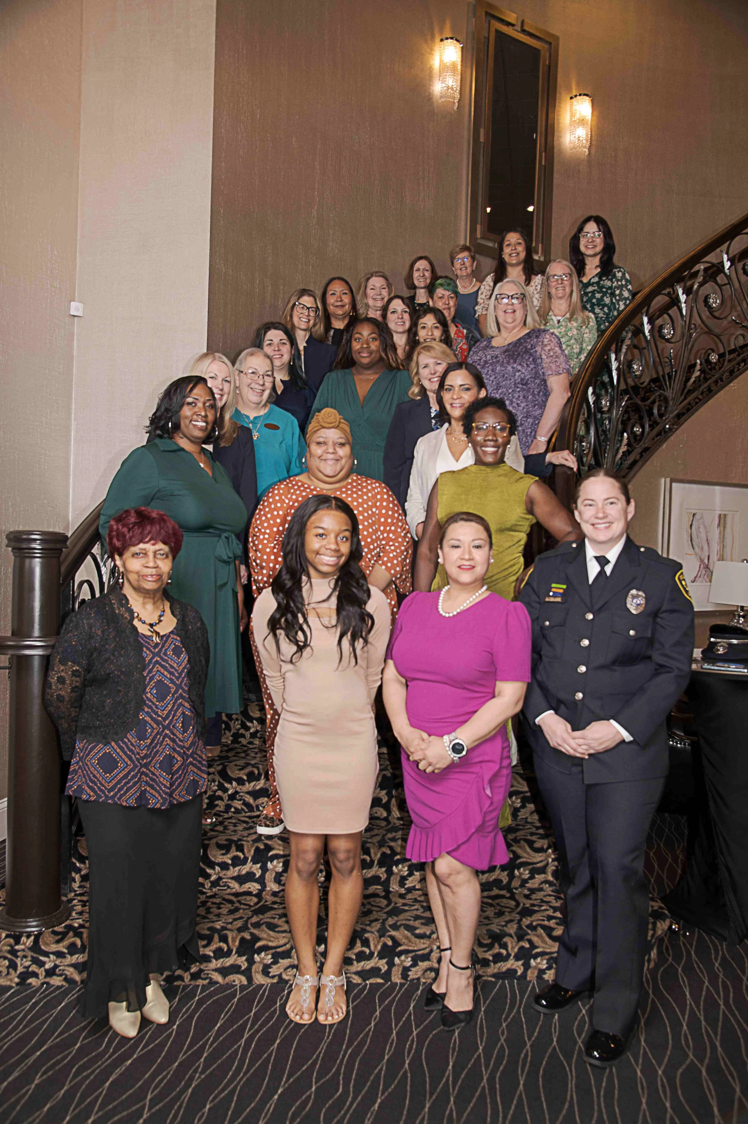 Group of women and one man posing on a staircase in a formal setting. The women are dressed in colorful dresses and blouses, and the man is in a police or military uniform. The background features a large mirror, warm lighting fixtures, and a pattern