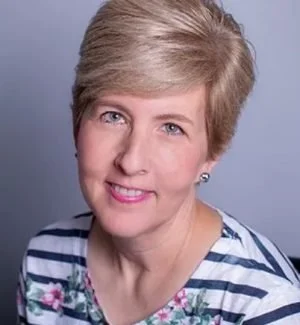 A woman with gray hair, glasses, and a patterned black-and-white scarf smiling against a blue cloudy background.