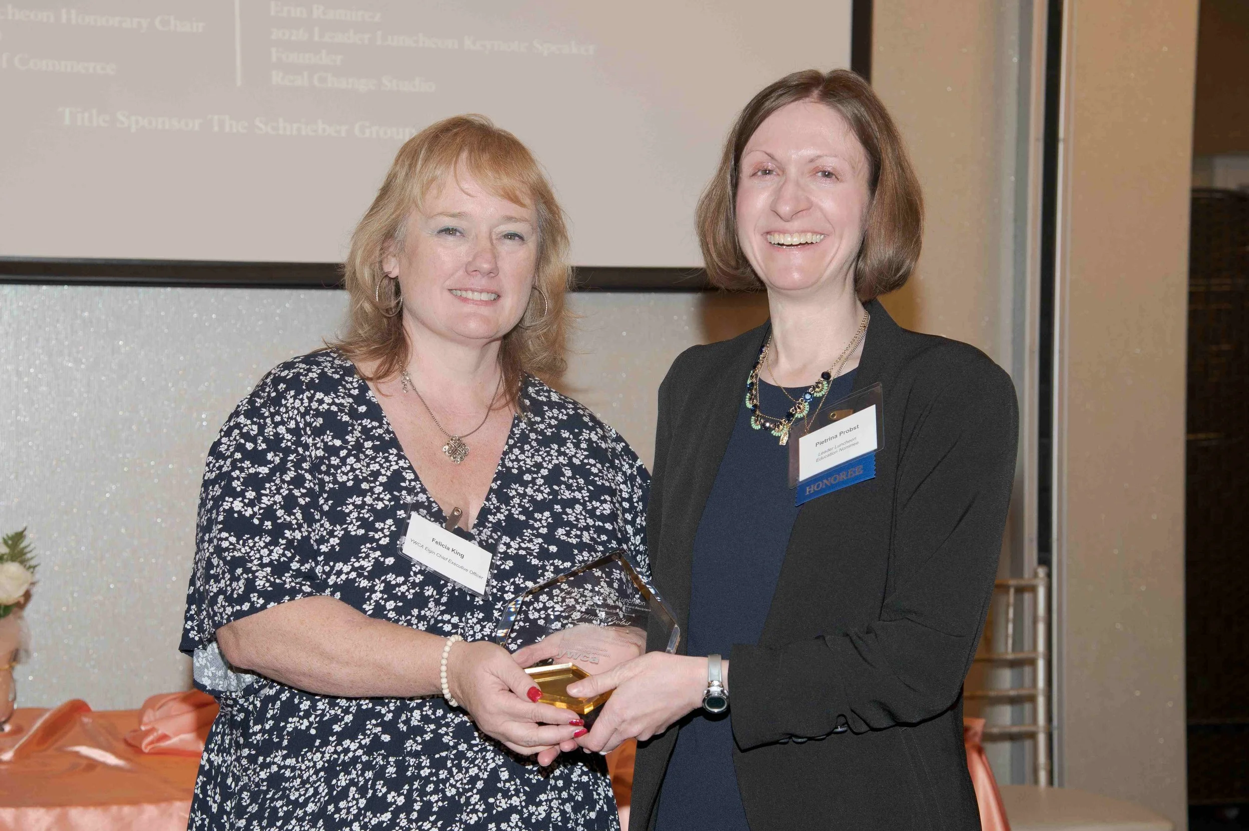 Two women standing side by side, smiling for the camera. The woman on the left has shoulder-length red hair and is wearing a black and white floral dress, holding a glass award. The woman on the right has short brown hair, is wearing a dark suit jack