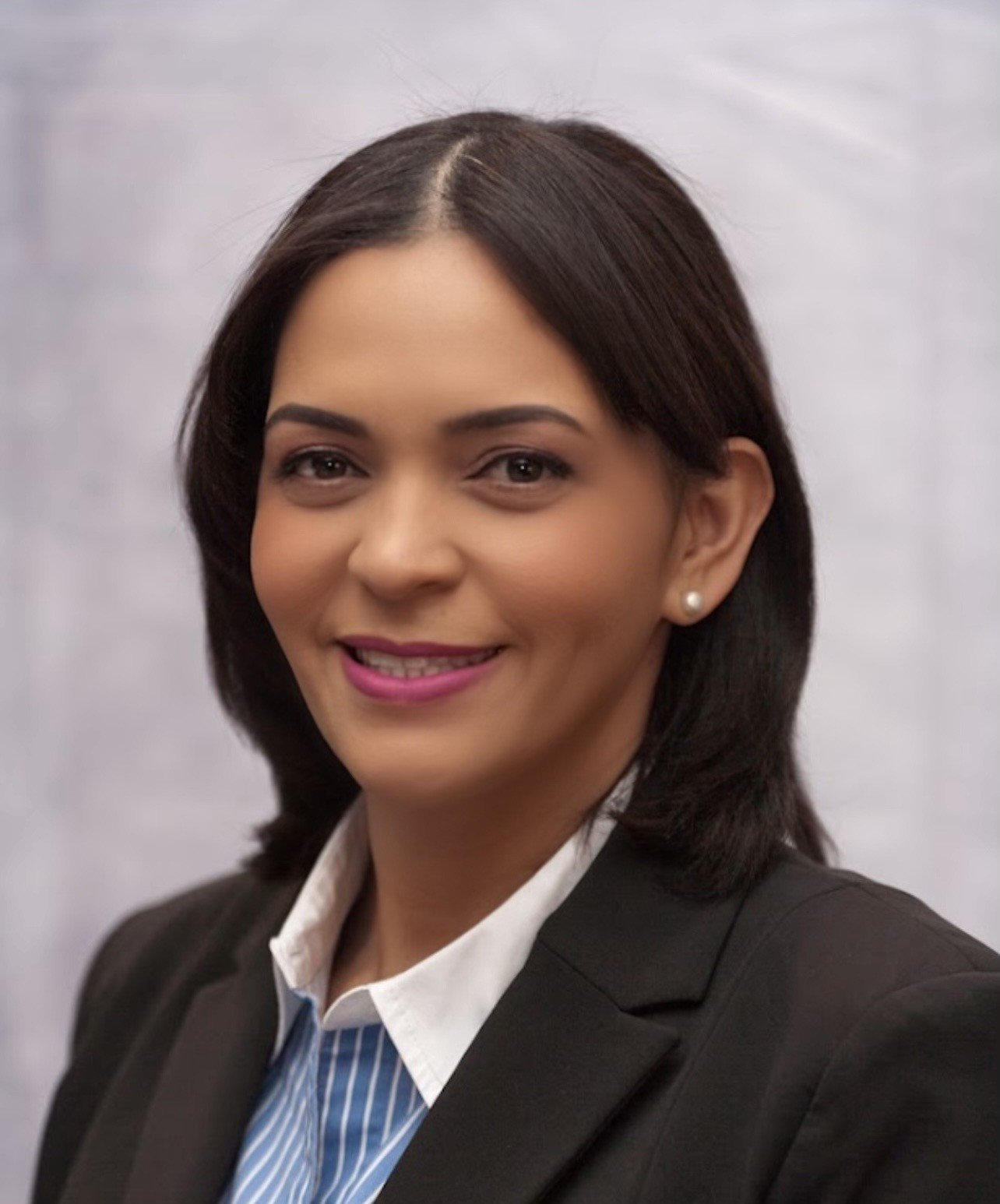 Professional woman with short brown hair and earrings, wearing a black blazer, smiling, against a gray background.