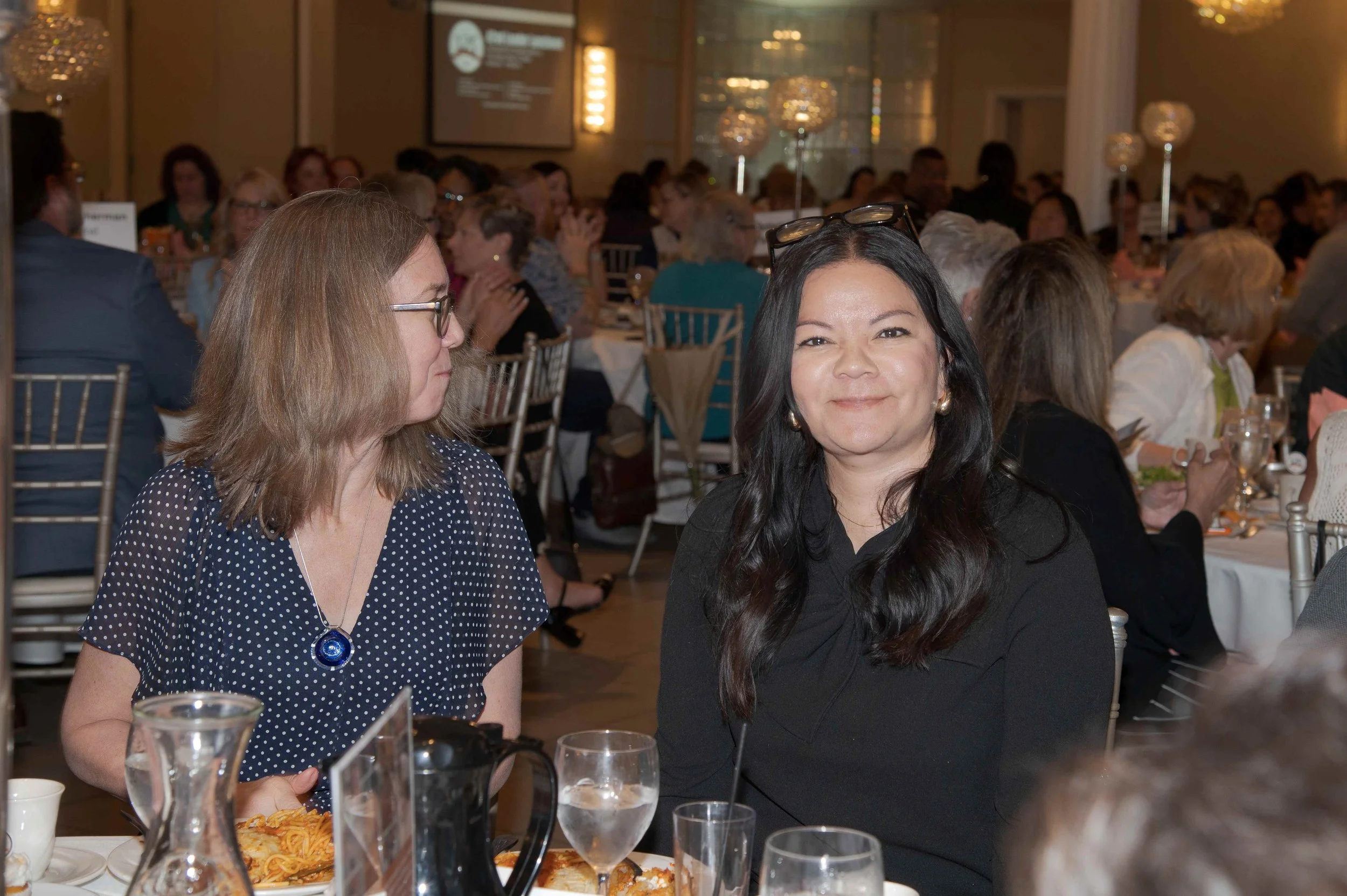 Two women sitting at a dinner table during a banquet or formal event, surrounded by many other guests seated at decorated tables in an elegant banquet hall.