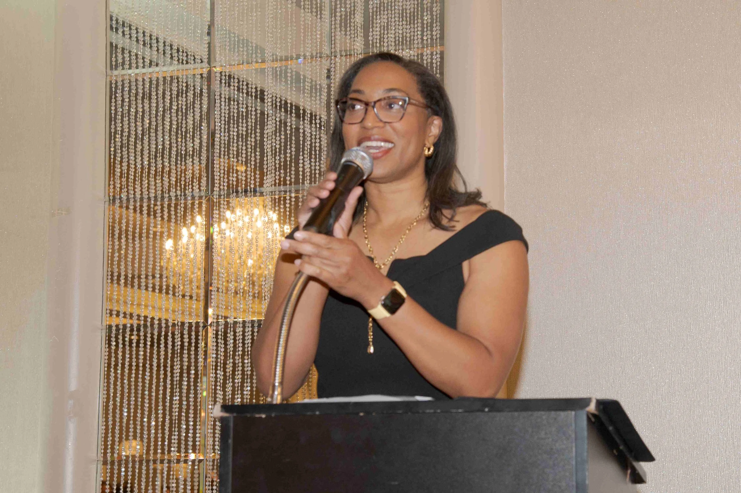 A woman with glasses and earrings speaking into a microphone at a formal event or speech, with a beaded curtain and light-colored wall in the background.