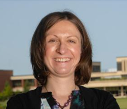 A woman in a red top and colorful skirt standing outdoors with arms crossed, smiling at the camera, with trees, water, and a bridge in the background.