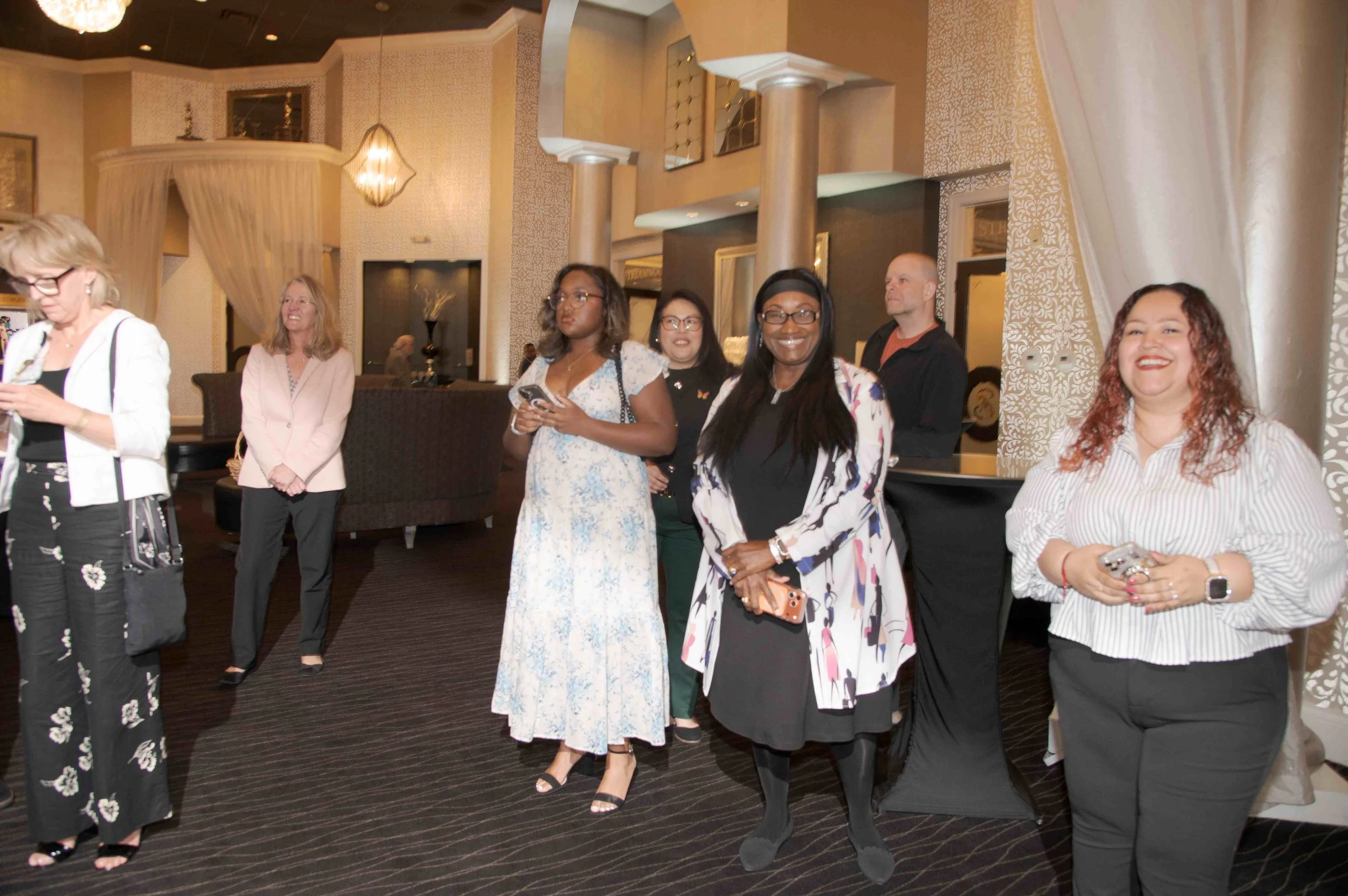A group of diverse women and a man standing in a well-lit, elegant indoor space, possibly at a social or networking event, with some women smiling and others looking serious. They are dressed in business or casual attire.