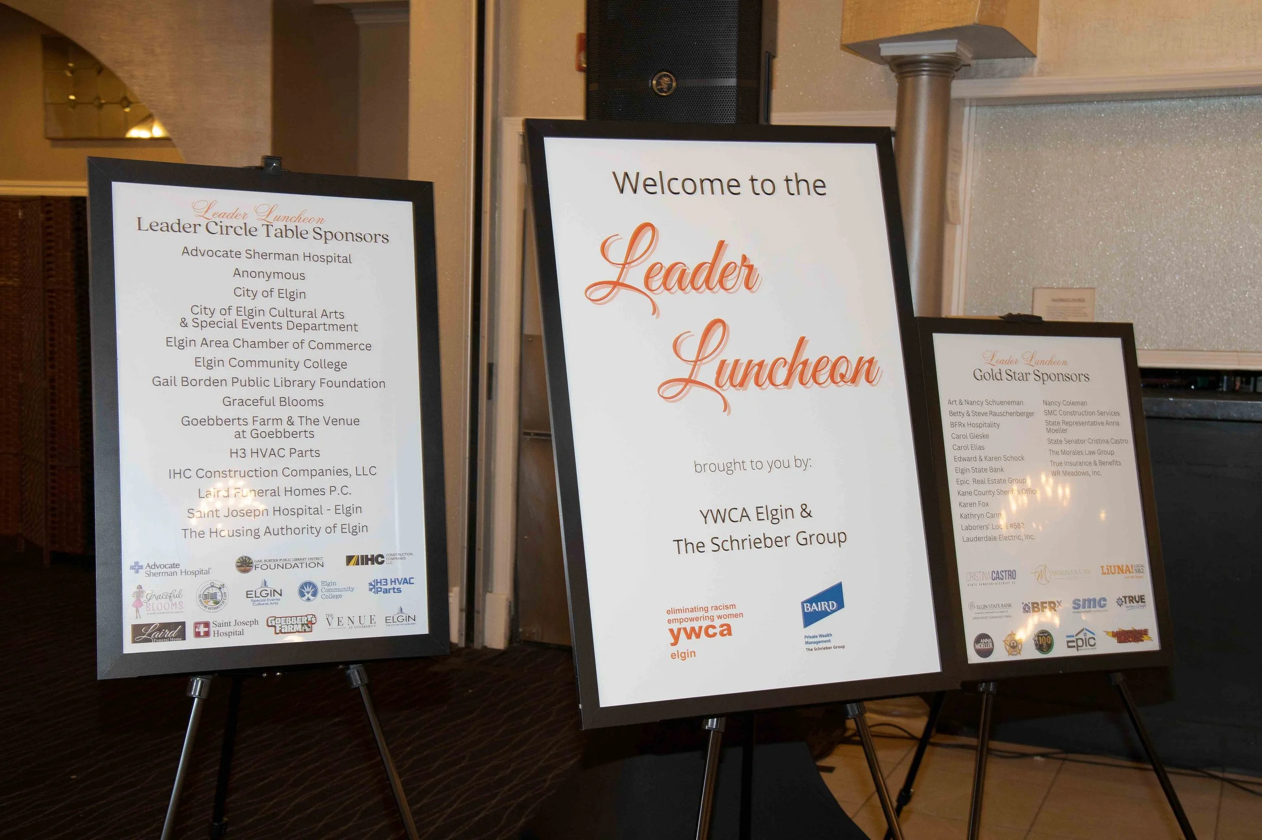 Three framed event programs on black easels at a conference, featuring sponsor and organization logos, with a beige wall and a speaker in the background.