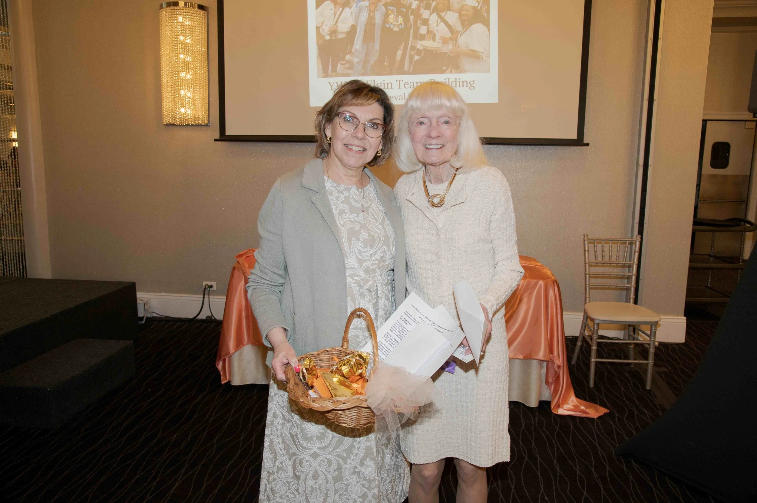 Two women stand together smiling in a room with a presentation screen and chairs behind them. One woman holds a basket of gold-wrapped chocolates, and the other holds papers.