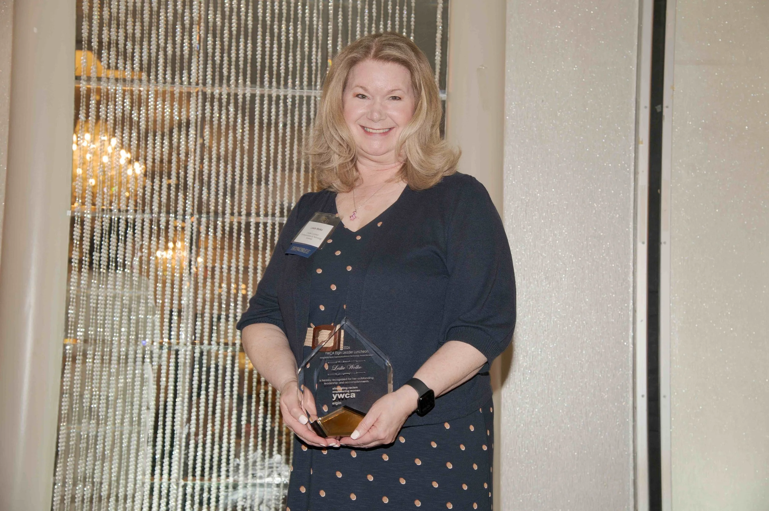 A woman with shoulder-length blonde hair wearing a navy blue dress with small peach-colored dots, holding an award plaque at the YWCA Elgin recognition event, standing in front of a beaded curtain and sparkly wall.