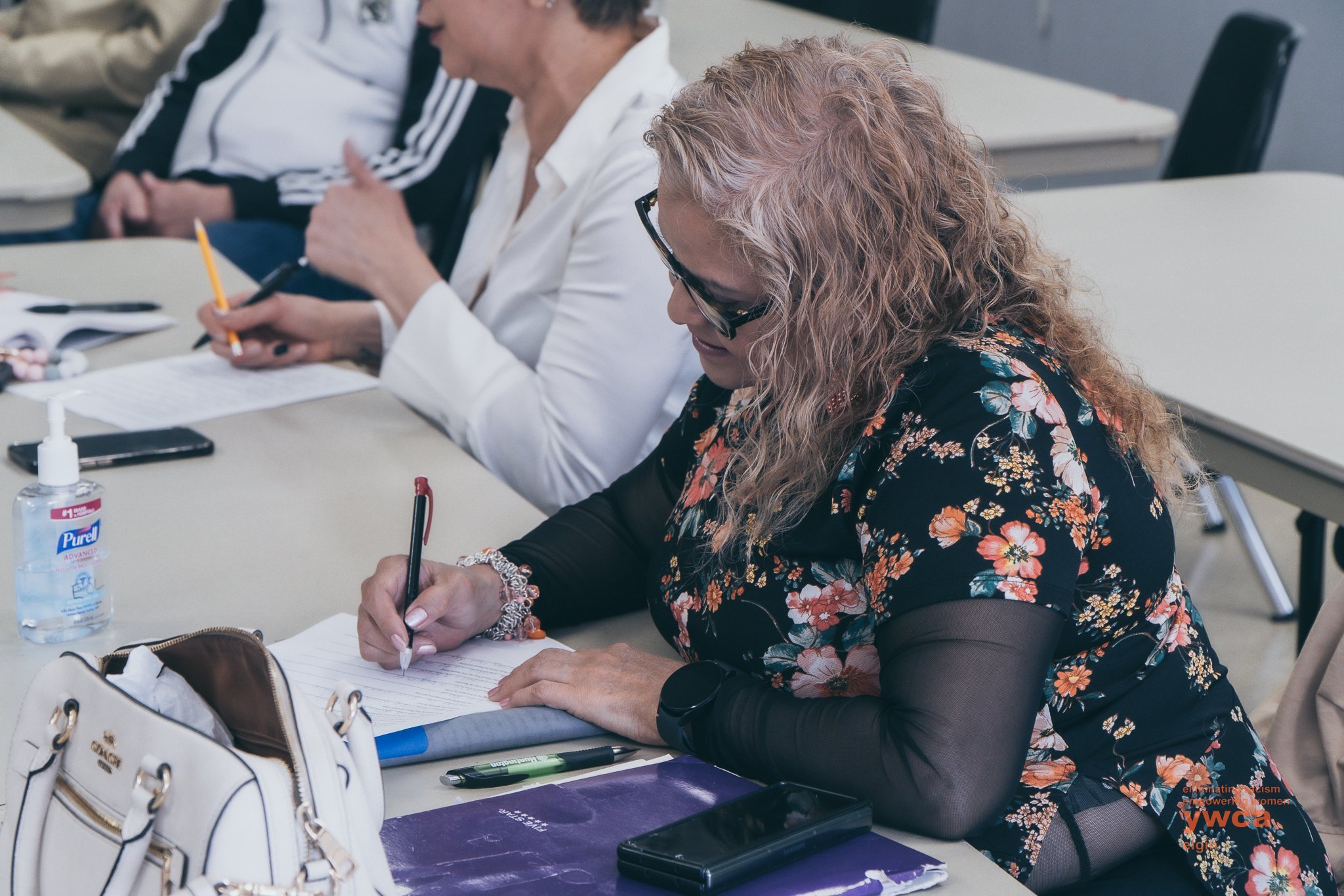 A woman with curly hair wearing glasses and a floral top taking notes during a meeting or classroom setting, with other people visible in the background.