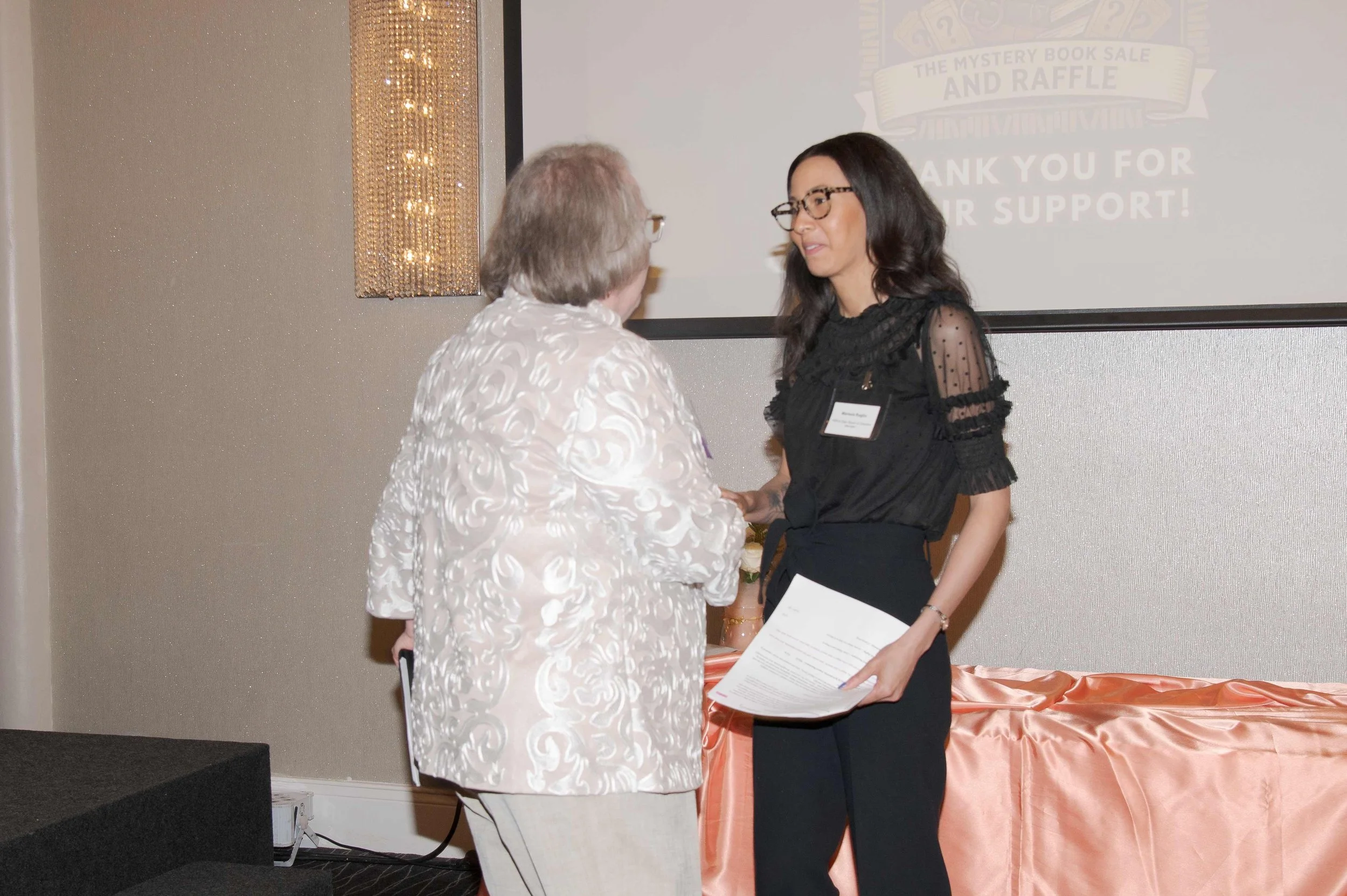 Two women shaking hands at a formal event, one older with gray hair and a patterned white jacket, the other younger with long dark hair wearing glasses, a black blouse with ruffled sleeves, and holding a document. A screen in the background displays 