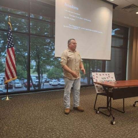 A man stands in front of a presentation screen in a conference room, with an American flag to his left and a table with a sign that says voting. There are large windows showing parked cars and trees outside.
