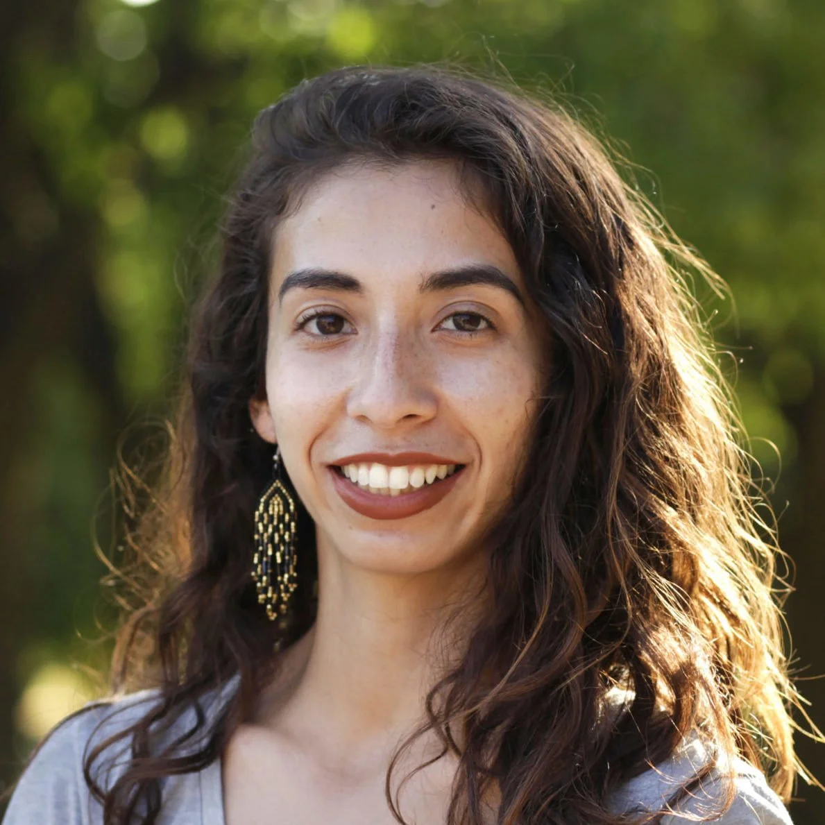 Portrait of a woman with dark brown hair, wearing a white collared shirt and a black graduation gown, against a blue background.