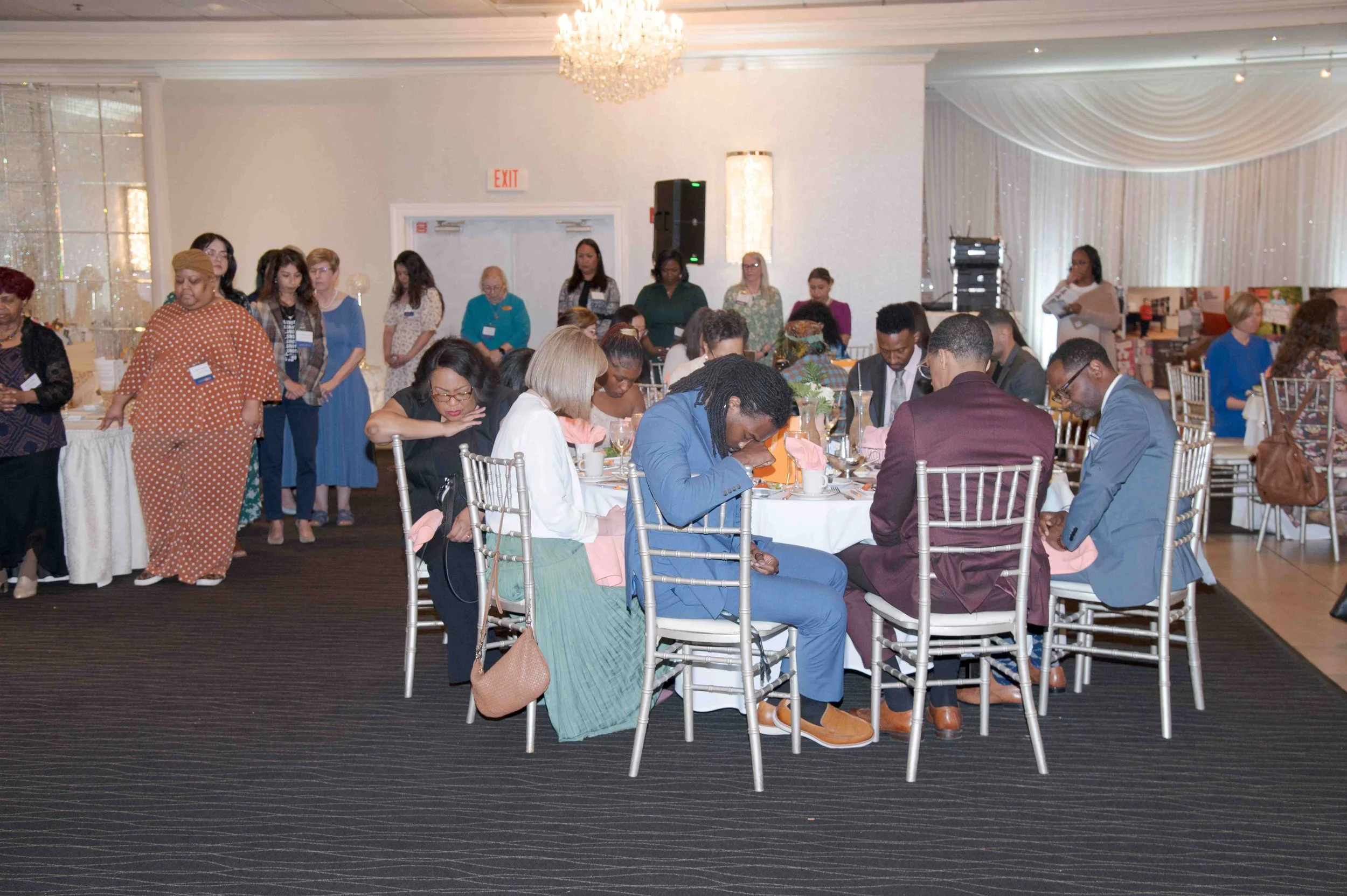 A group of people participating in a prayer circle at a formal event, with attendees kneeling and eyes closed around a decorated table in a banquet hall with chandeliers.