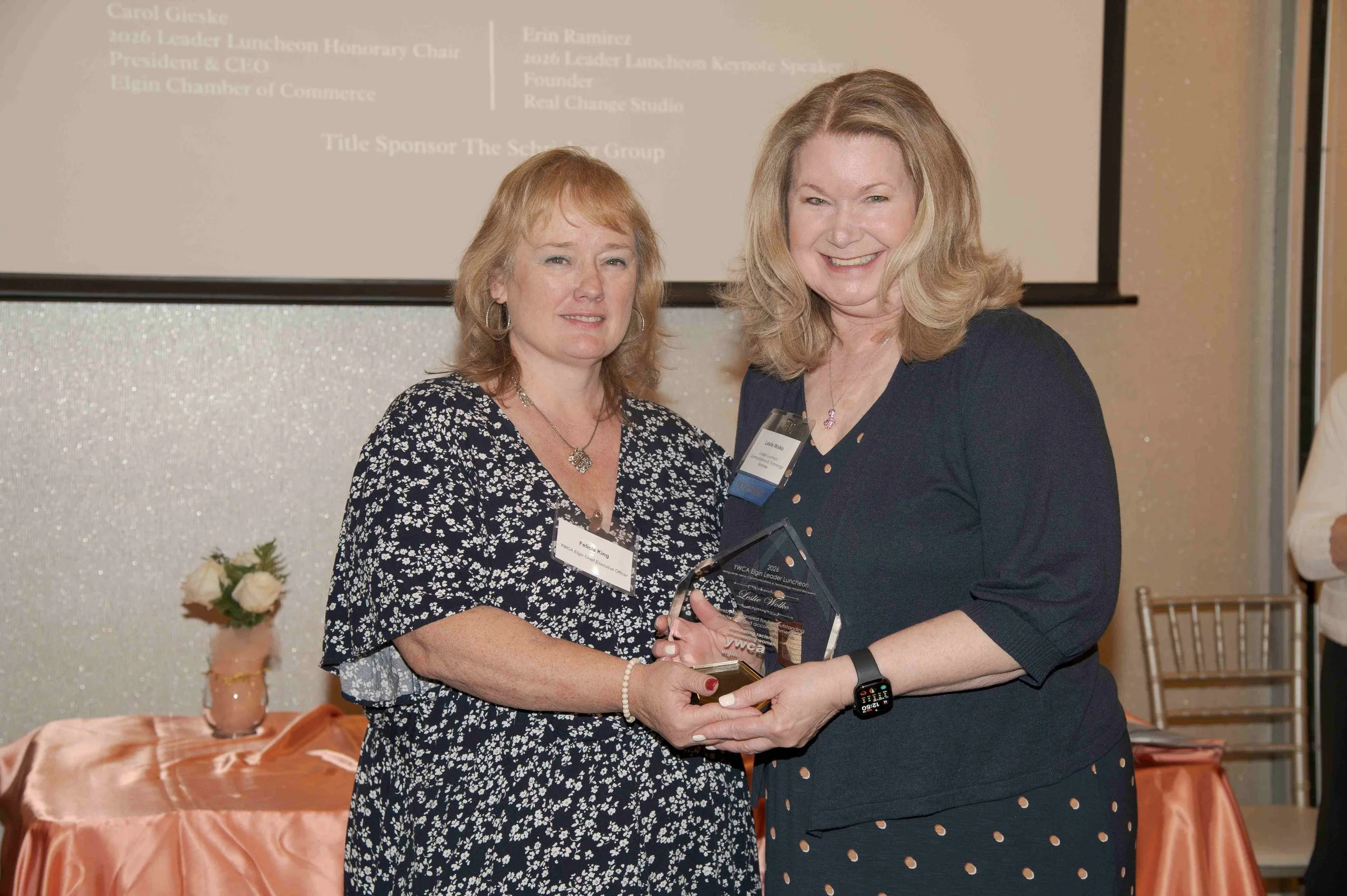 Two women are standing together at an event, smiling and shaking hands. The woman on the left is holding a glass award and wearing a dark floral dress. The woman on the right is wearing a dark jacket with a blue dress that has white polka dots. They 