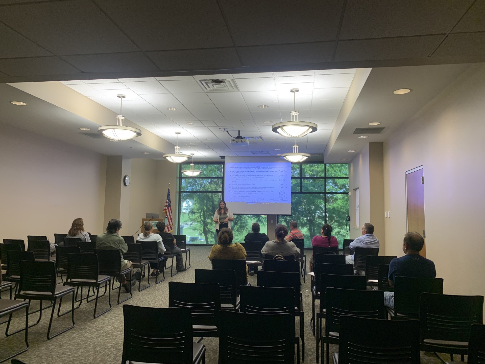 A woman standing in front of an audience during a presentation in a conference room with large windows, a projector screen, and an American flag.