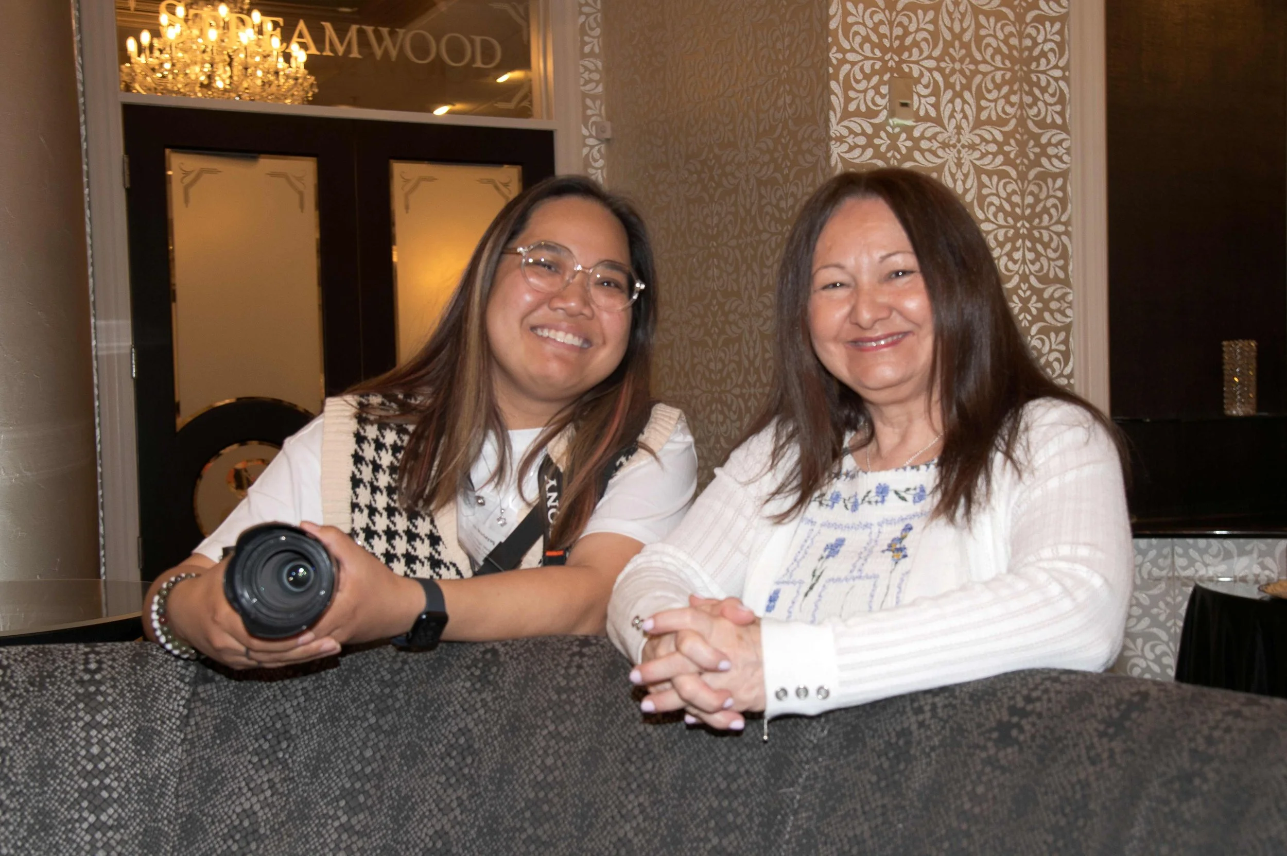 Two women sit at a table, smiling, with a chandelier and decorative wallpaper in the background.