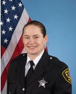 Photograph of a woman with wavy gray hair, glasses, a patterned scarf, and a smile, set against a blue cloudy background.