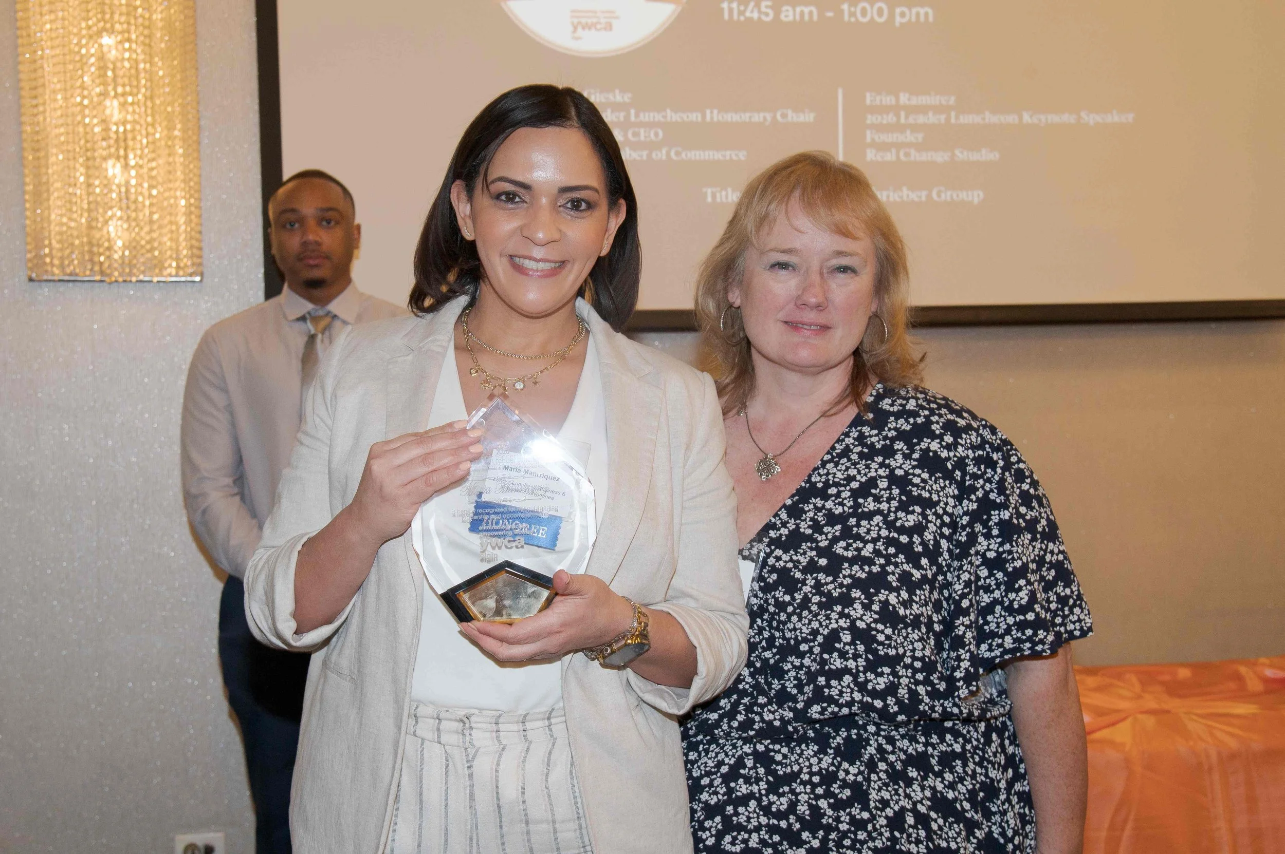Two women smiling at an award ceremony, with a man in the background. One woman holds a glass award. They are dressed in business casual and appear to be at a formal event.