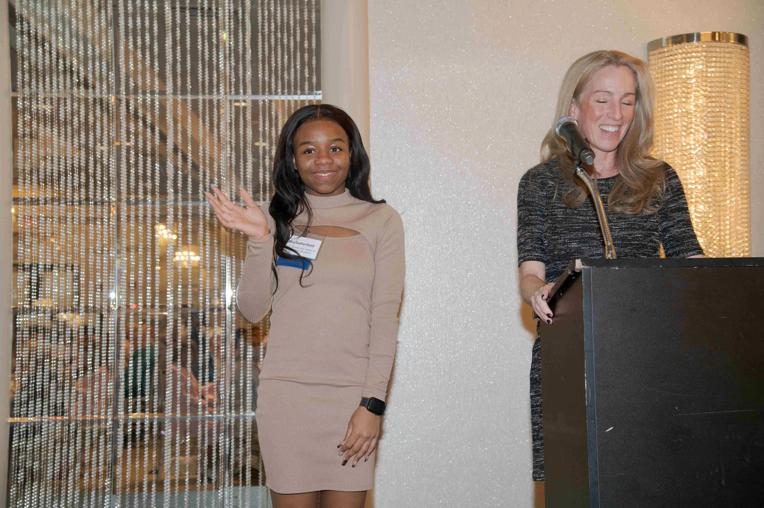 A woman with long blonde hair is speaking at a podium, smiling, wearing a black dress. Next to her, a woman with long dark hair in a beige dress waves and smiles, standing in front of a beaded curtain.