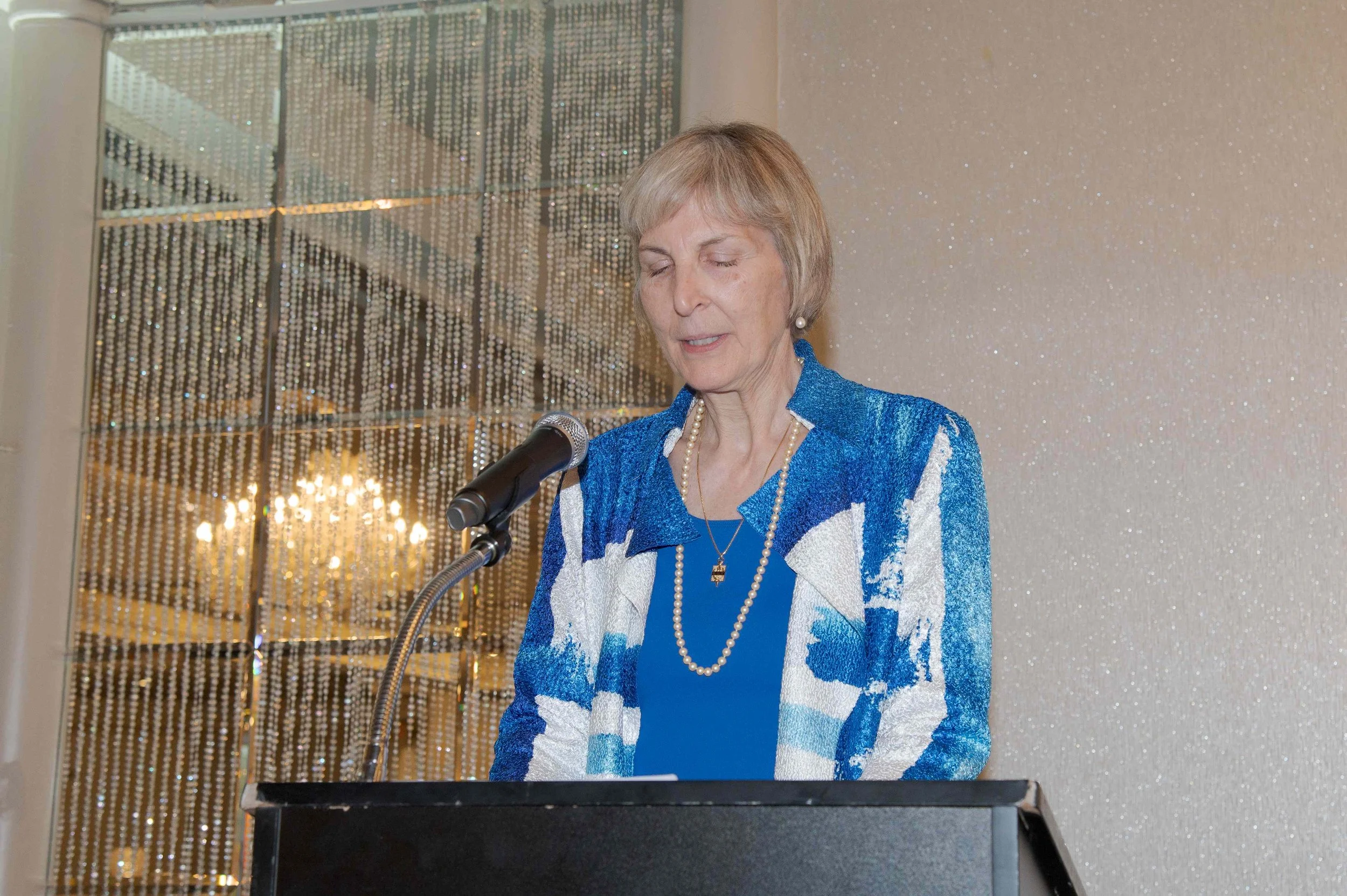 An elderly woman with short gray hair wearing a blue and white patterned jacket, pearl earrings, and a pendant necklace, standing at a podium with a microphone, with a glittery wall and a mirrored textured background behind her.