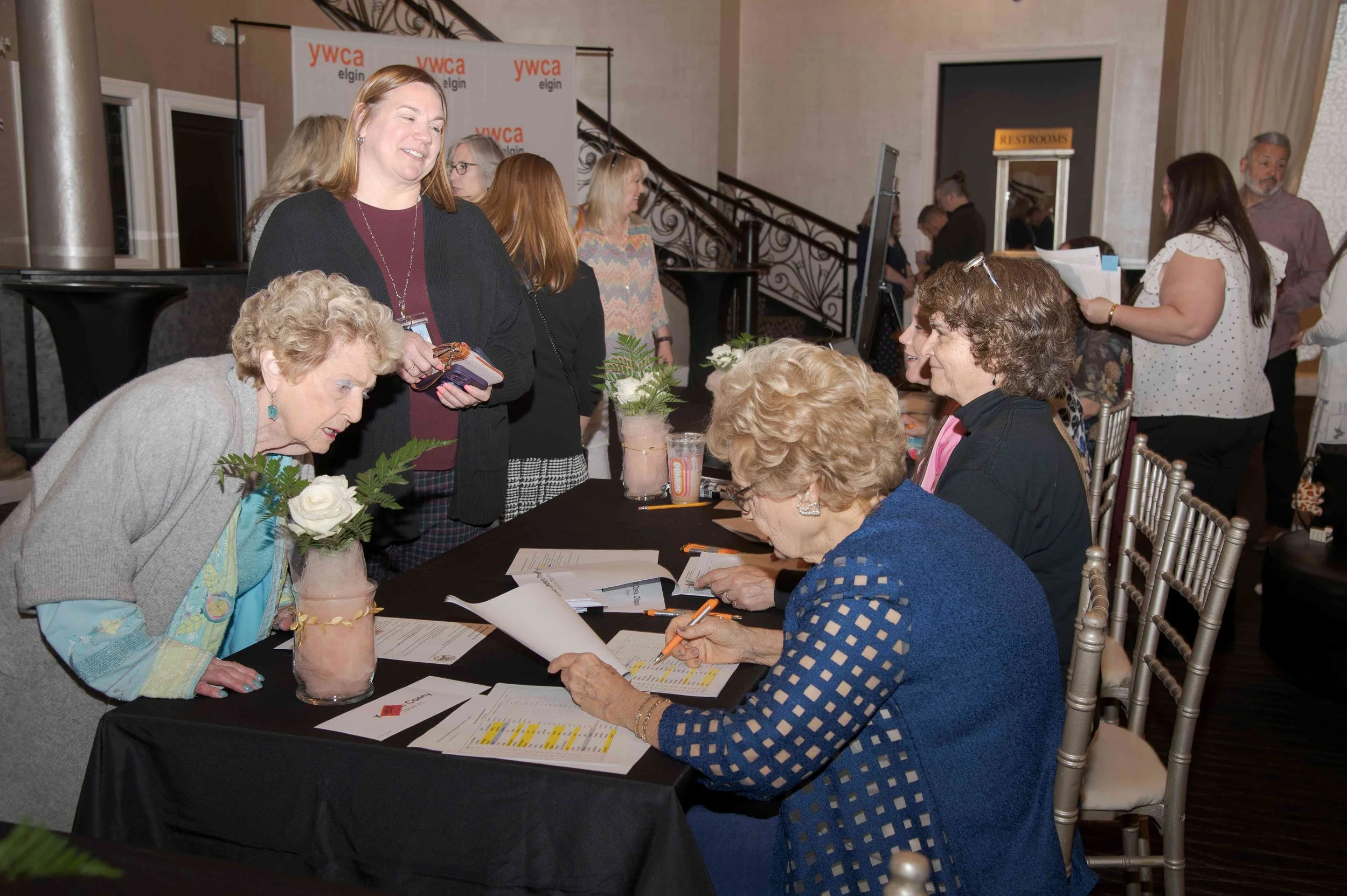 Group of women at a registration or sign-in table at a conference or event, with some signing documents and others engaged in conversation, inside a decorated venue with a staircase and a YMCA banner in the background.