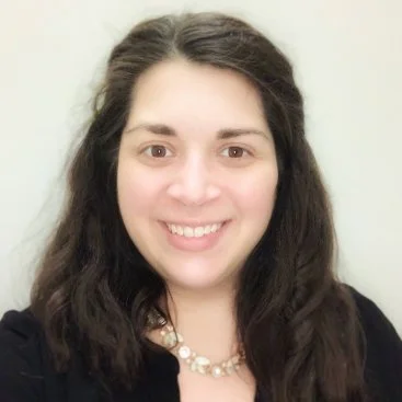 A woman with shoulder-length brown hair, blue eyes, wearing hoop earrings and a black blazer, smiling at the camera.