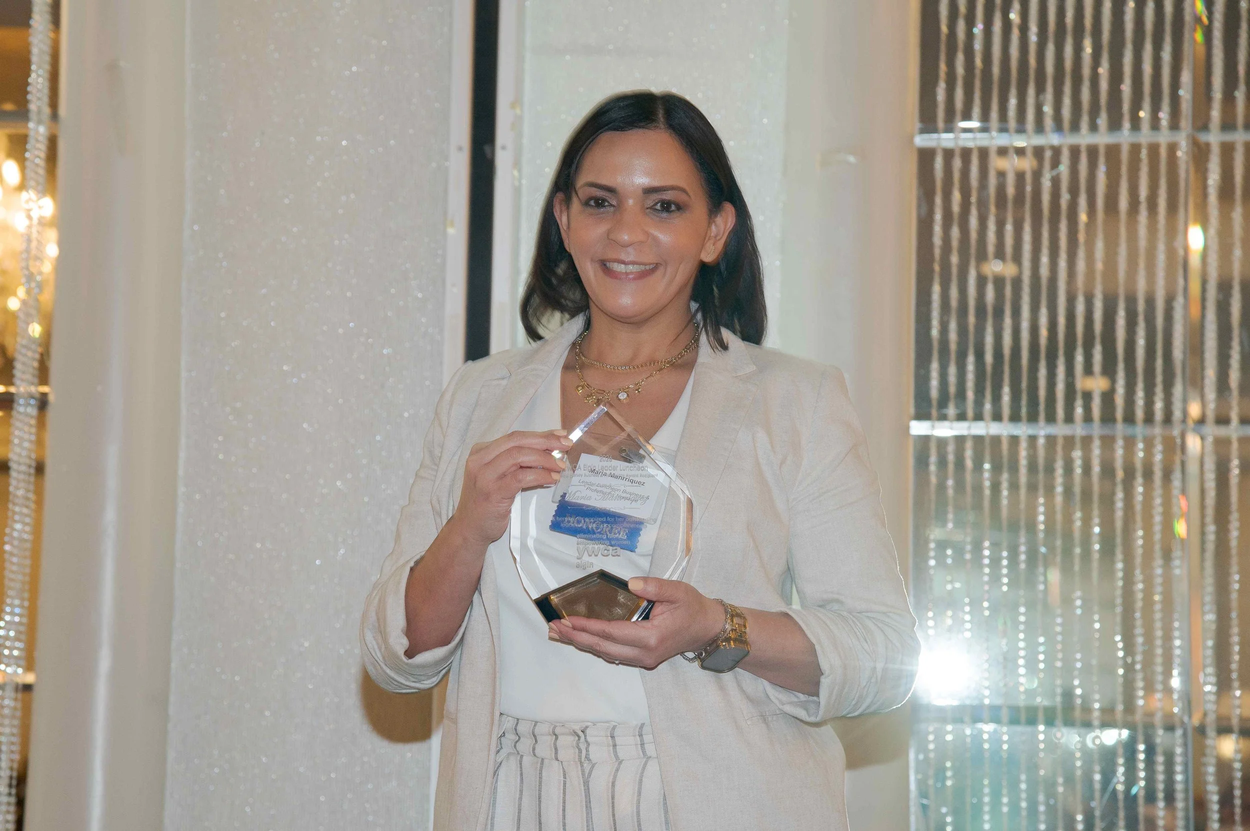 A woman with dark hair holding an award or plaque, standing in front of a sparkly curtain and silver beads.