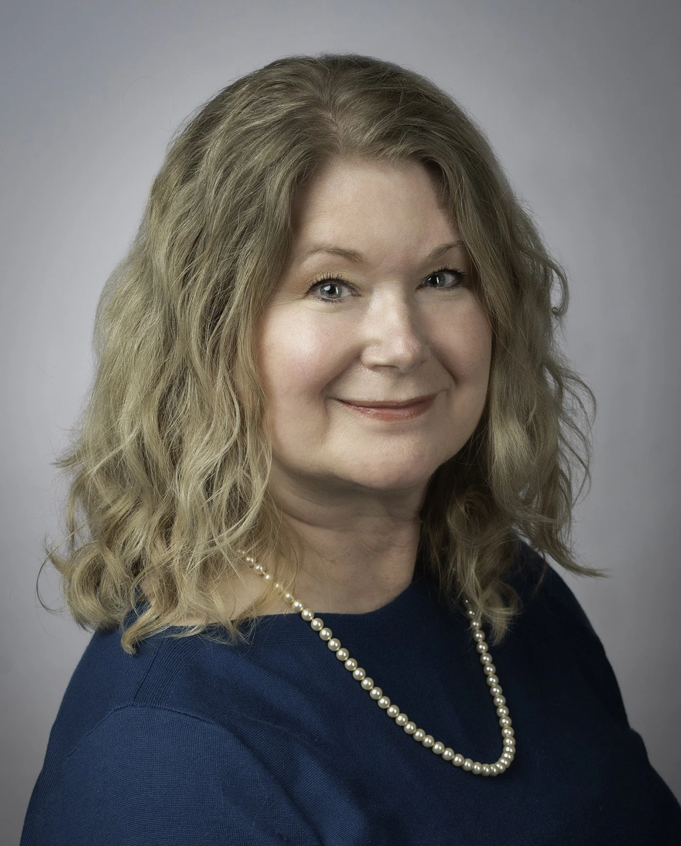 A woman with blonde hair and glasses, wearing a black blazer, smiling against a gray background.