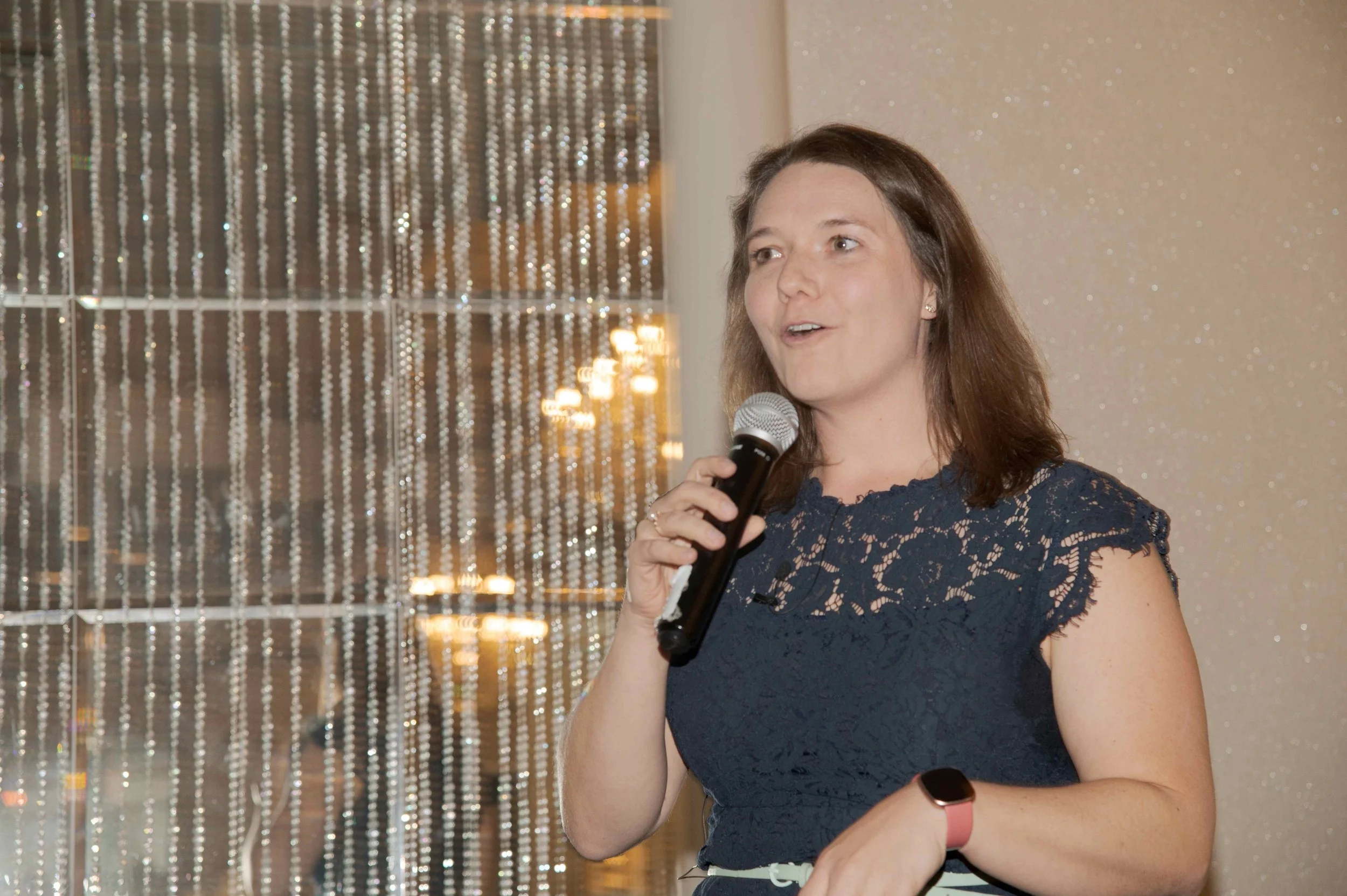 A woman with shoulder-length brown hair wearing a navy lace dress, holding a microphone and speaking at an event with a sparkling beige background and beaded curtain.