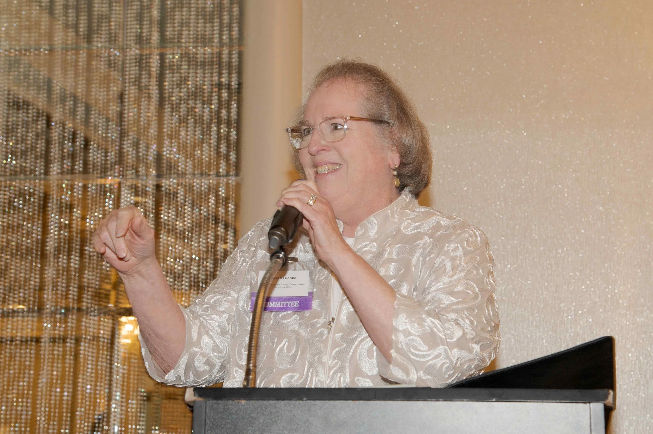 An older woman with glasses and earrings speaking into a microphone at a conference, standing behind a podium with a purple badge reading 'COMMITTEE'.
