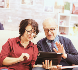 An elderly man and a woman are sitting together looking at a tablet, smiling and engaging with the device.