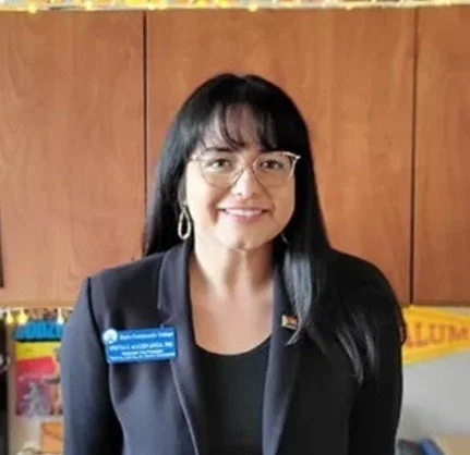 A woman with short blond hair, blue eyes, and light skin smiling, wearing a denim jacket and a purple and pink scarf, against a soft gray background.