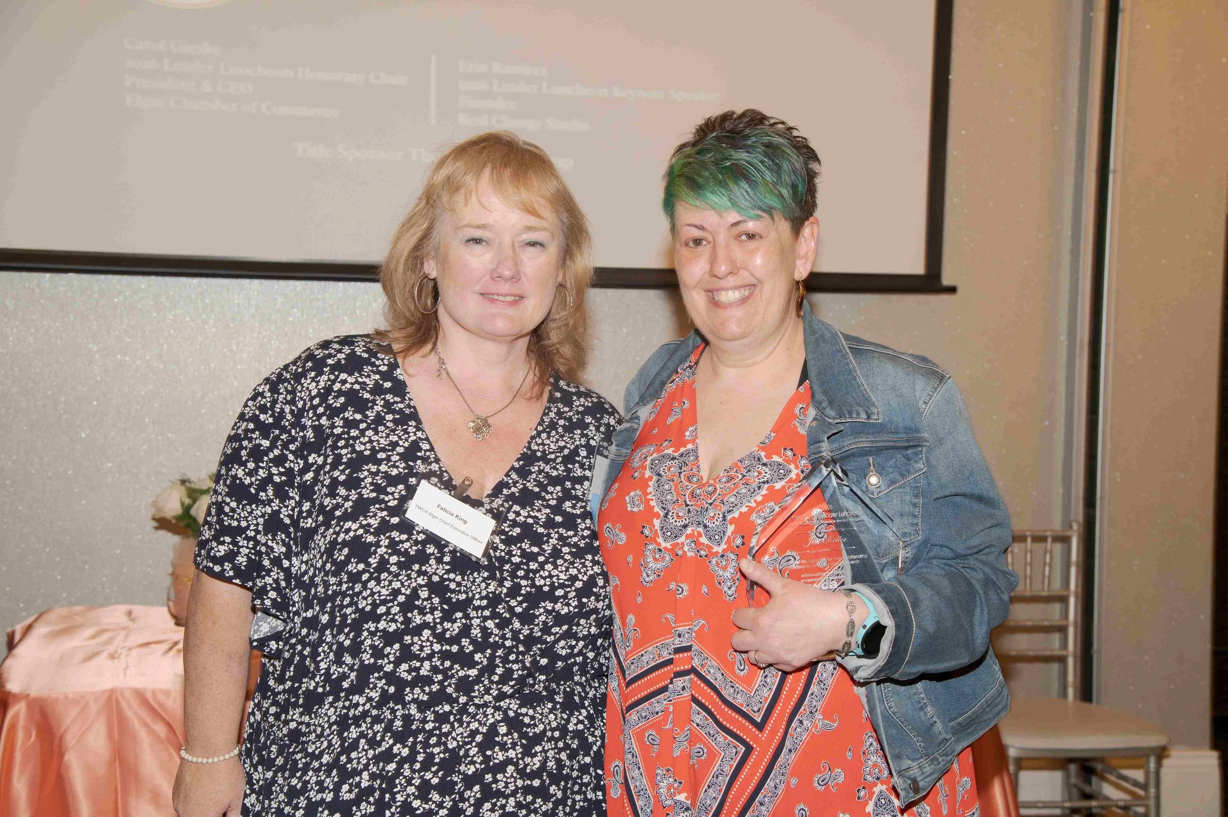 Two women standing together at an event, one wearing a black and white floral dress with a name tag and the other wearing an orange and navy paisley dress with a denim jacket, both smiling at the camera.