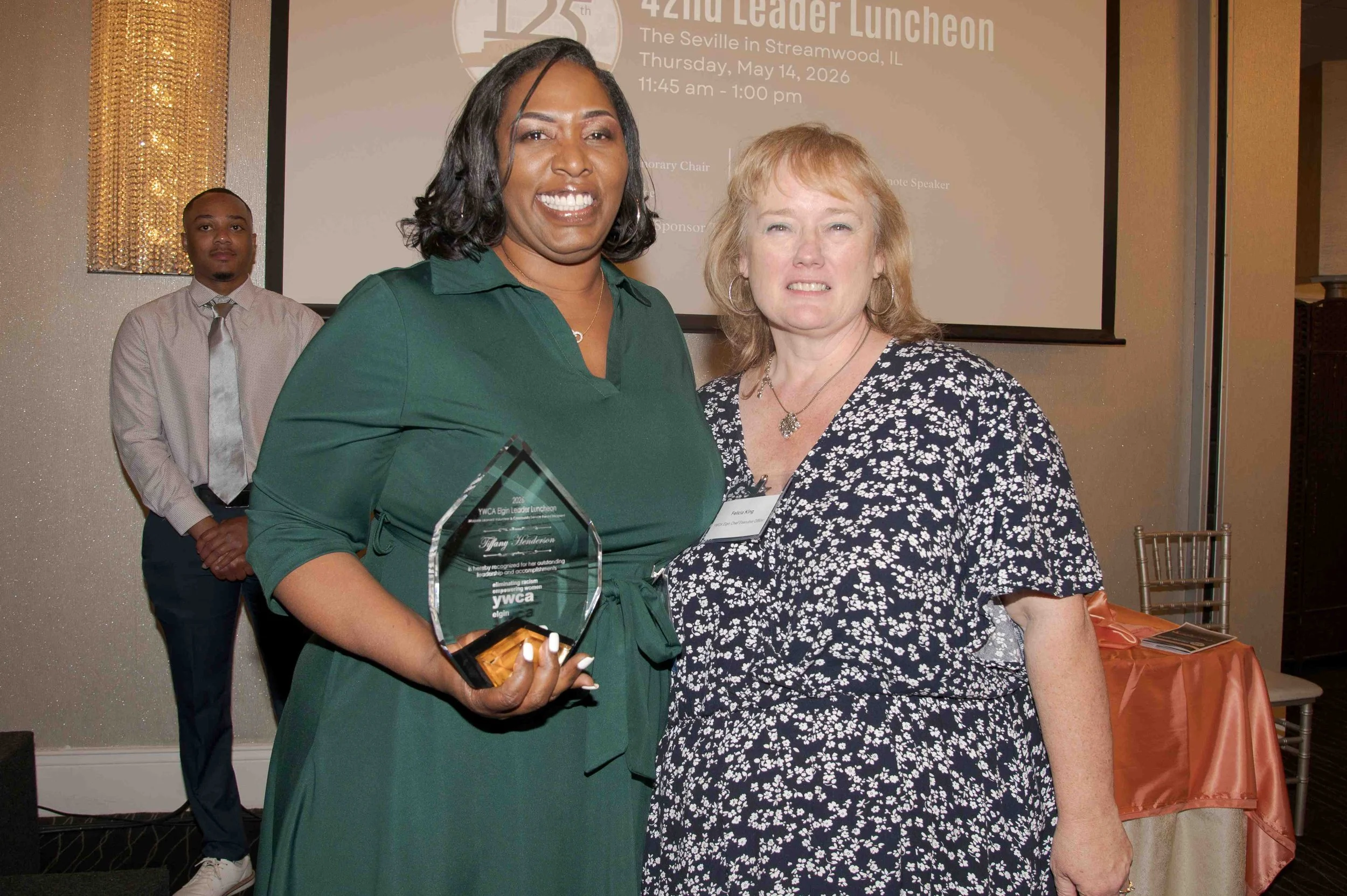 Two women stand together, one holding a glass award, at an awards ceremony with a presentation screen in the background displaying event details. The woman on the left is wearing a green dress, and the woman on the right is wearing a black and white 