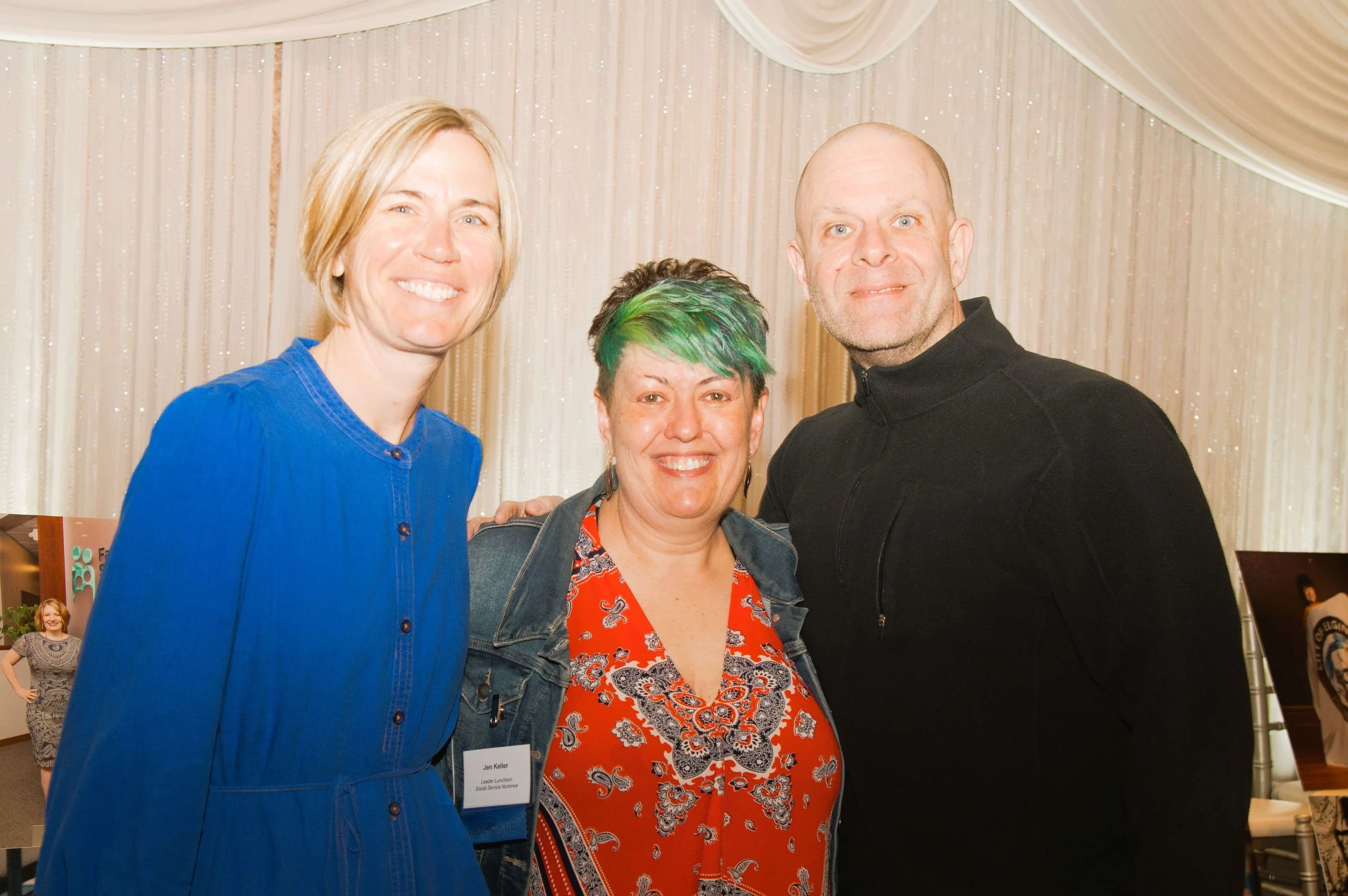Four smiling people standing close together with a beige curtain background. One woman has short blonde hair and is wearing a blue shirt. The second woman has short hair with green highlights and is wearing a denim jacket over an orange shirt with a 