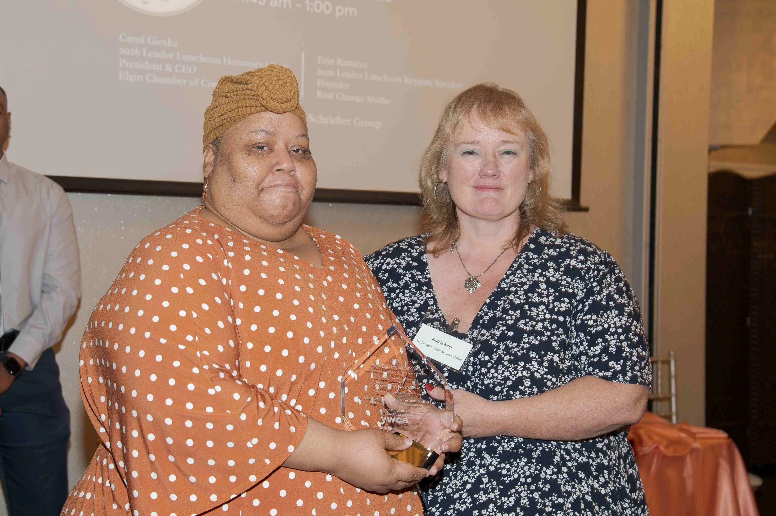 Two women standing together, one holding an award, at an indoor event. The woman on the left is wearing an orange polka dot dress and a brown headwrap. The woman on the right is wearing a black and white floral dress with earrings and a necklace. The